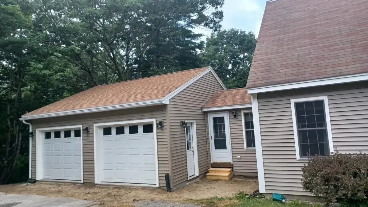 White two-car garage and beige house exterior with dormer windows and wooded yard