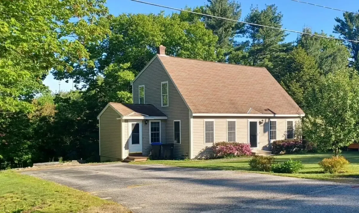 Small beige house with a tan roof, green shutters, and a gravel driveway surrounded by trees and shrubs