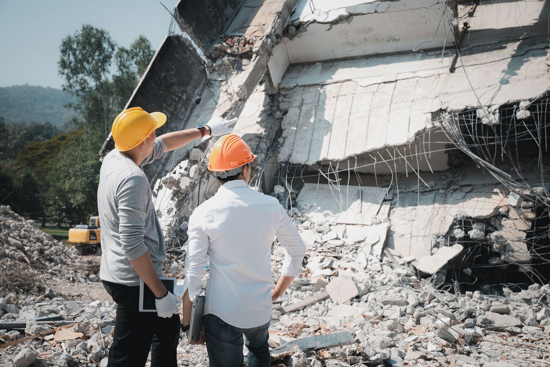 Two workers in hard hats inspect a collapsed concrete structure with rubble at the base.