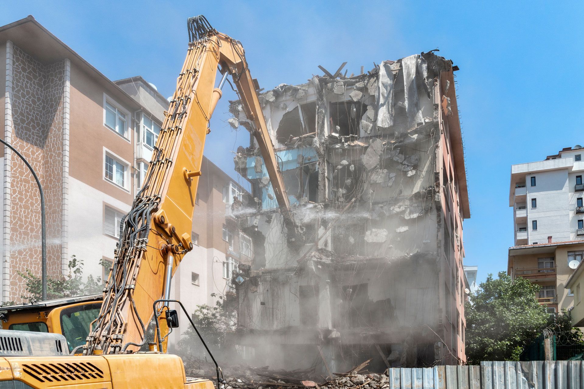 Yellow excavator demolishing a concrete building, with dust cloud in a residential area