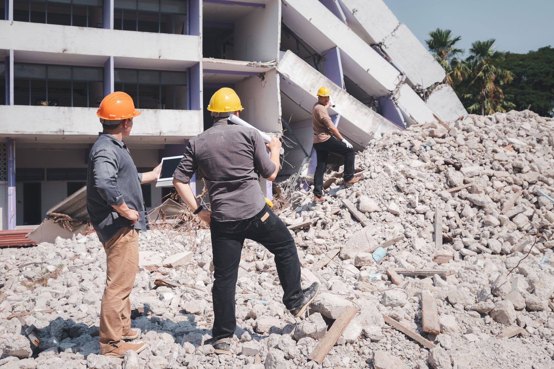 Workers in hard hats inspect rubble beside a damaged, partially collapsed building.