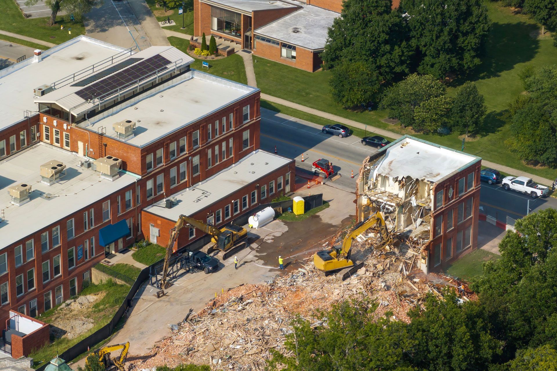 Aerial view of a building partially demolished, with rubble, construction equipment, and nearby brick buildings.