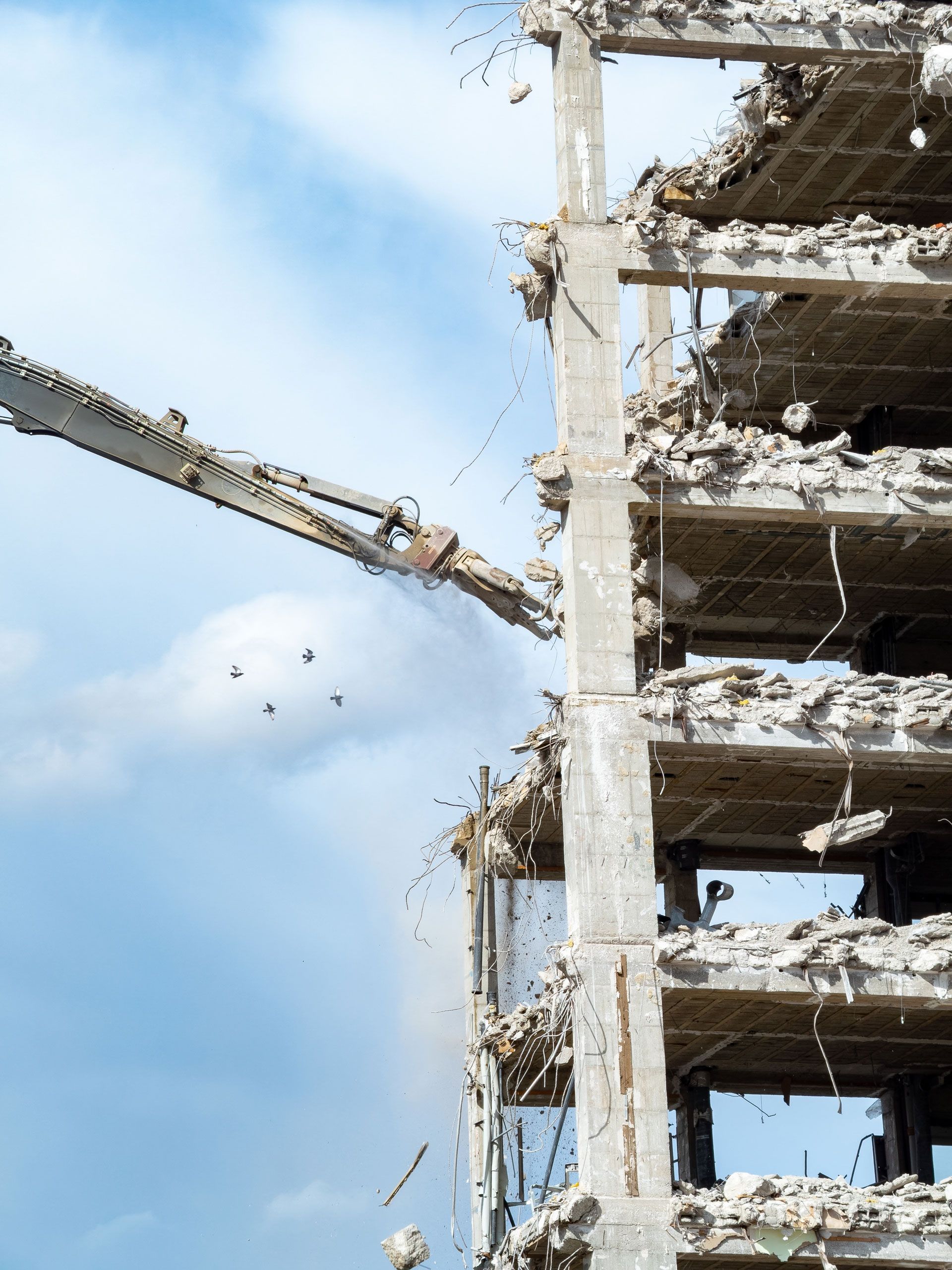 Demolition excavator arm tearing down a concrete building against a blue sky