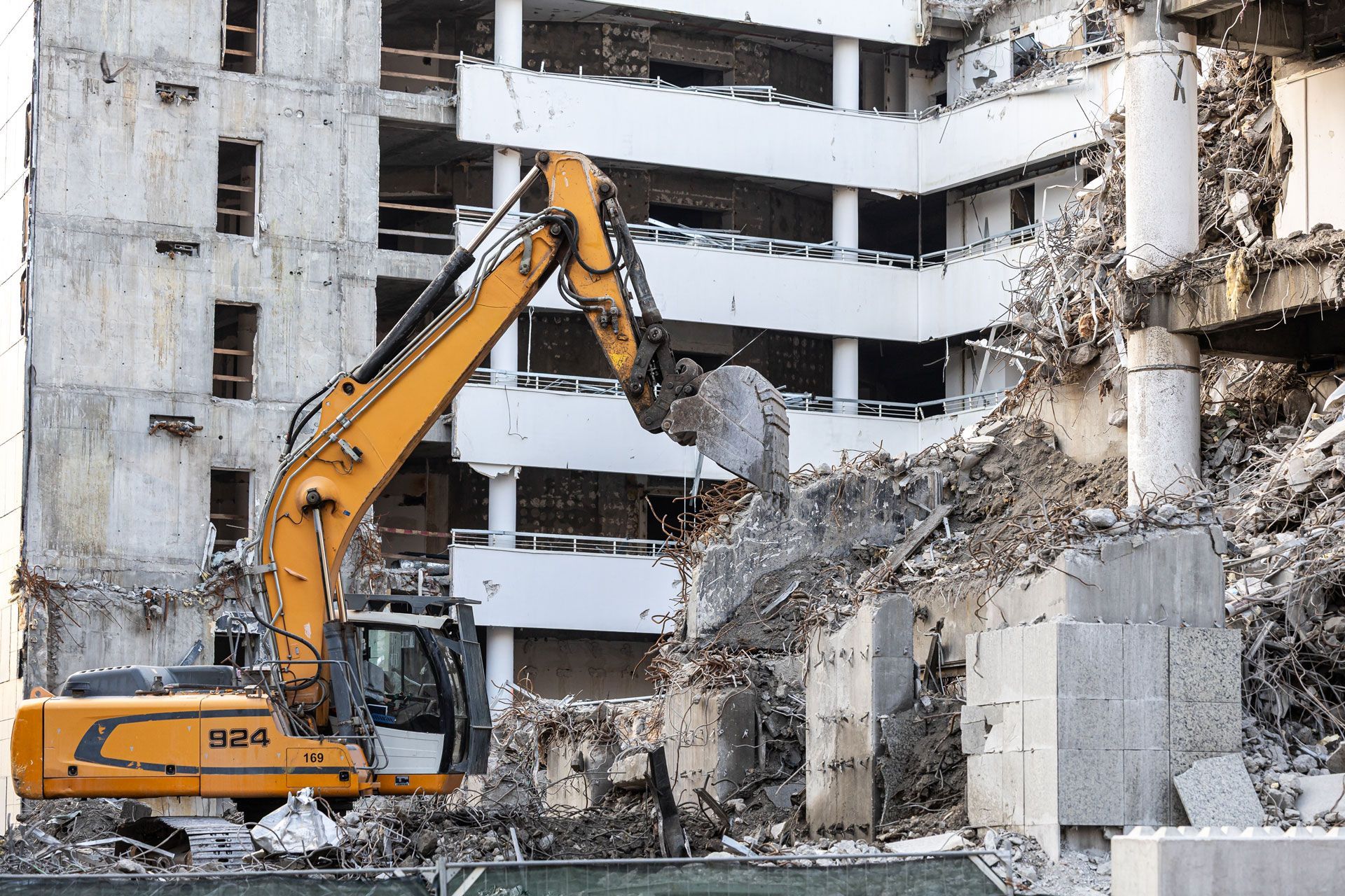 Excavator demolishing a damaged concrete building, with rubble and broken walls around it.
