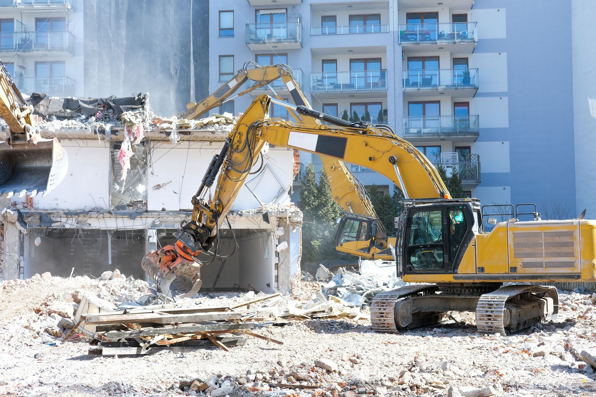 Yellow excavator demolishing a building, with rubble piled in front of an apartment block