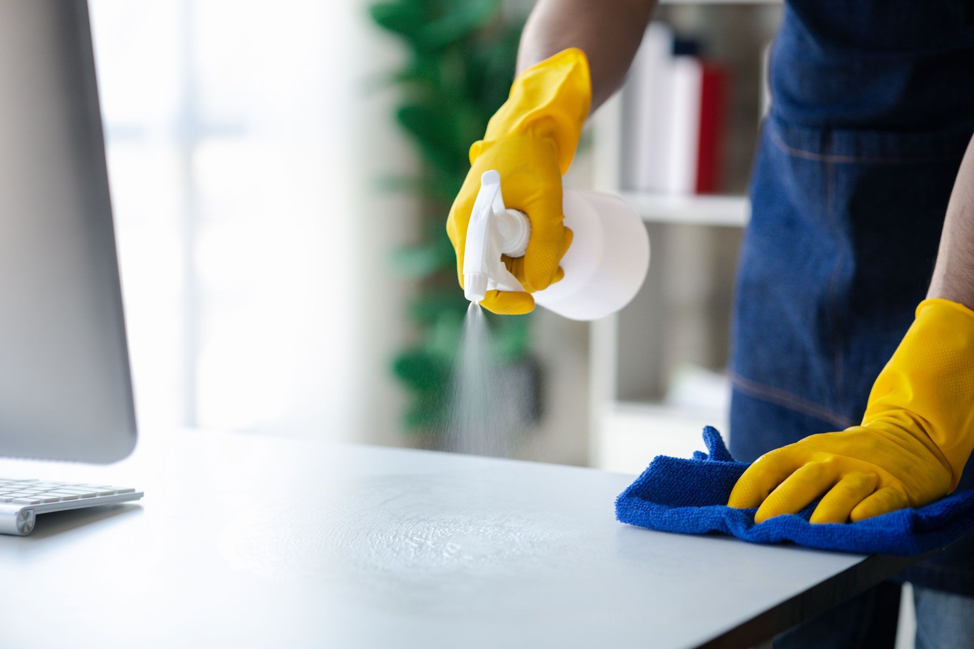 Person in yellow gloves cleaning a white desk with a blue cloth and spray bottle