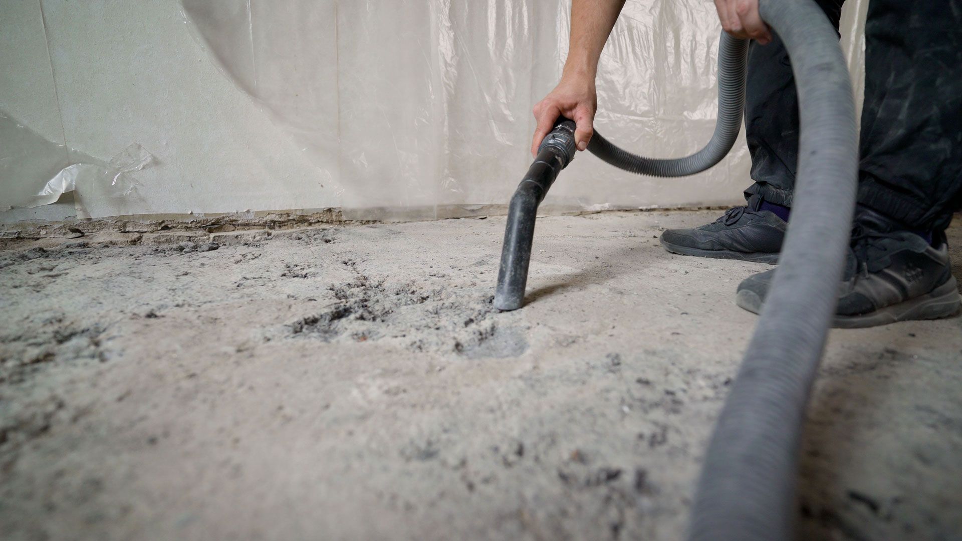 Worker using a vacuum hose to clean dust from a rough concrete floor indoors