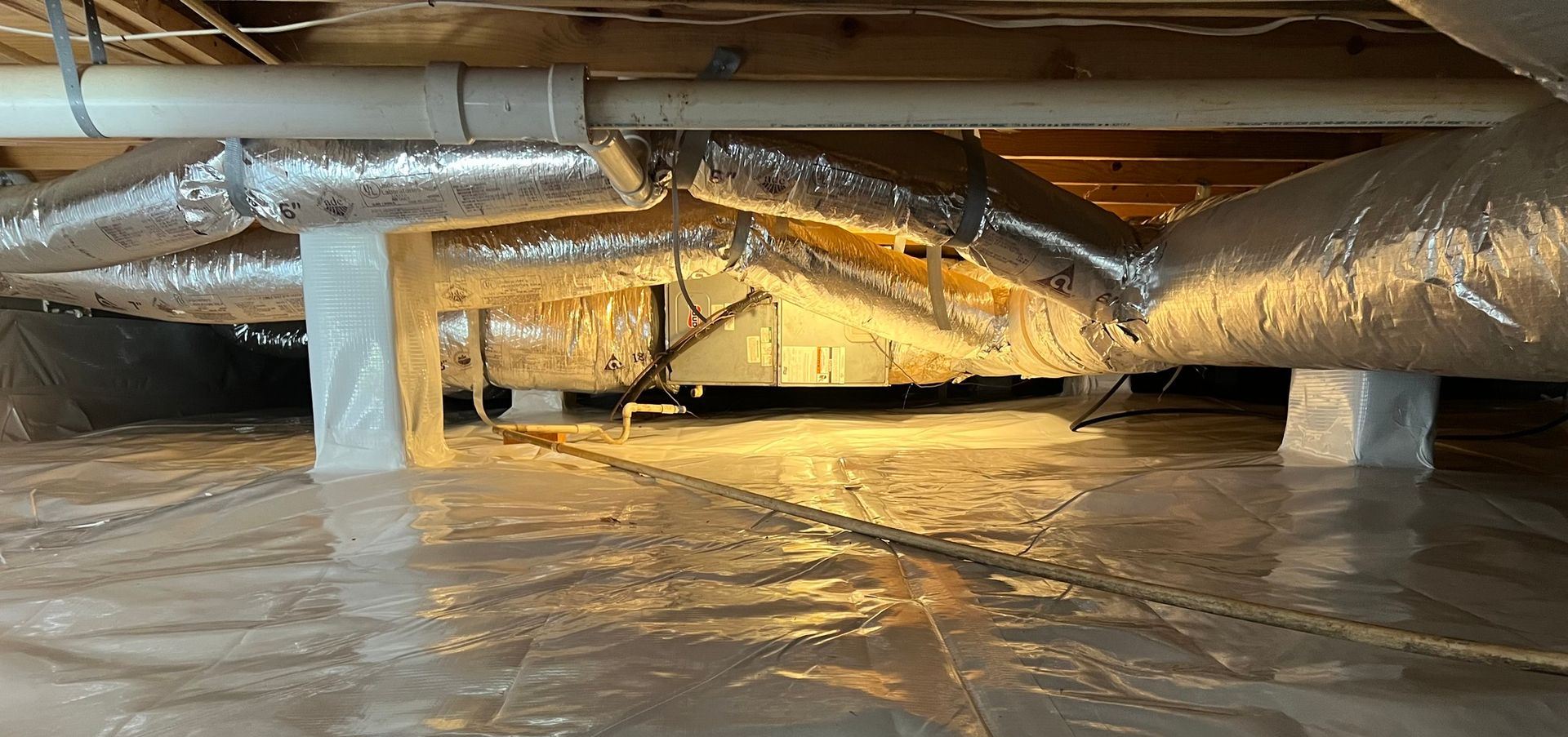 Low attic crawlspace with insulated ducts, support posts, and plastic-covered floor under warm light
