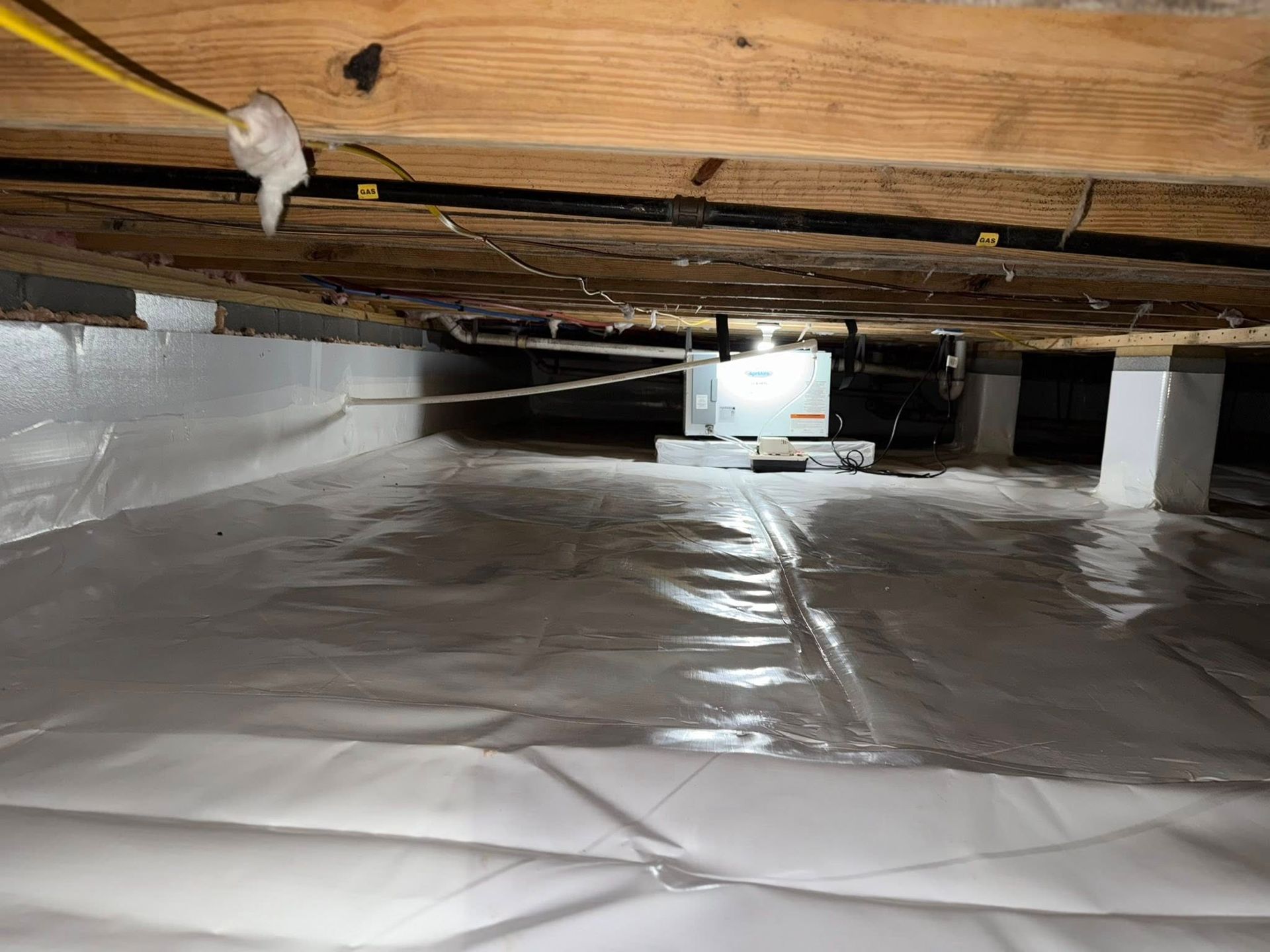 Crawlspace with white vapor barrier covering the ground. Wood beams overhead and a metal box in the distance.