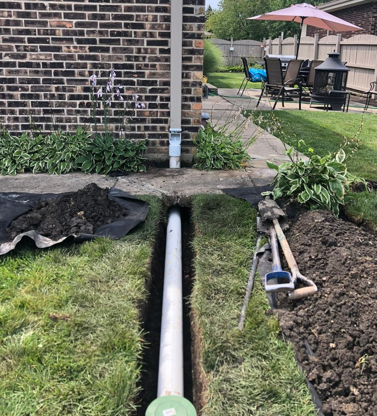 Backyard trench with a white pipe running from a brick house to a garden area; tools and patio in background