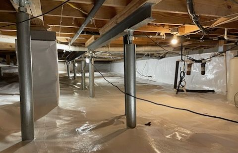 Crawl space with supports, white plastic sheeting, electrical wiring, and insulation against a white wall.
