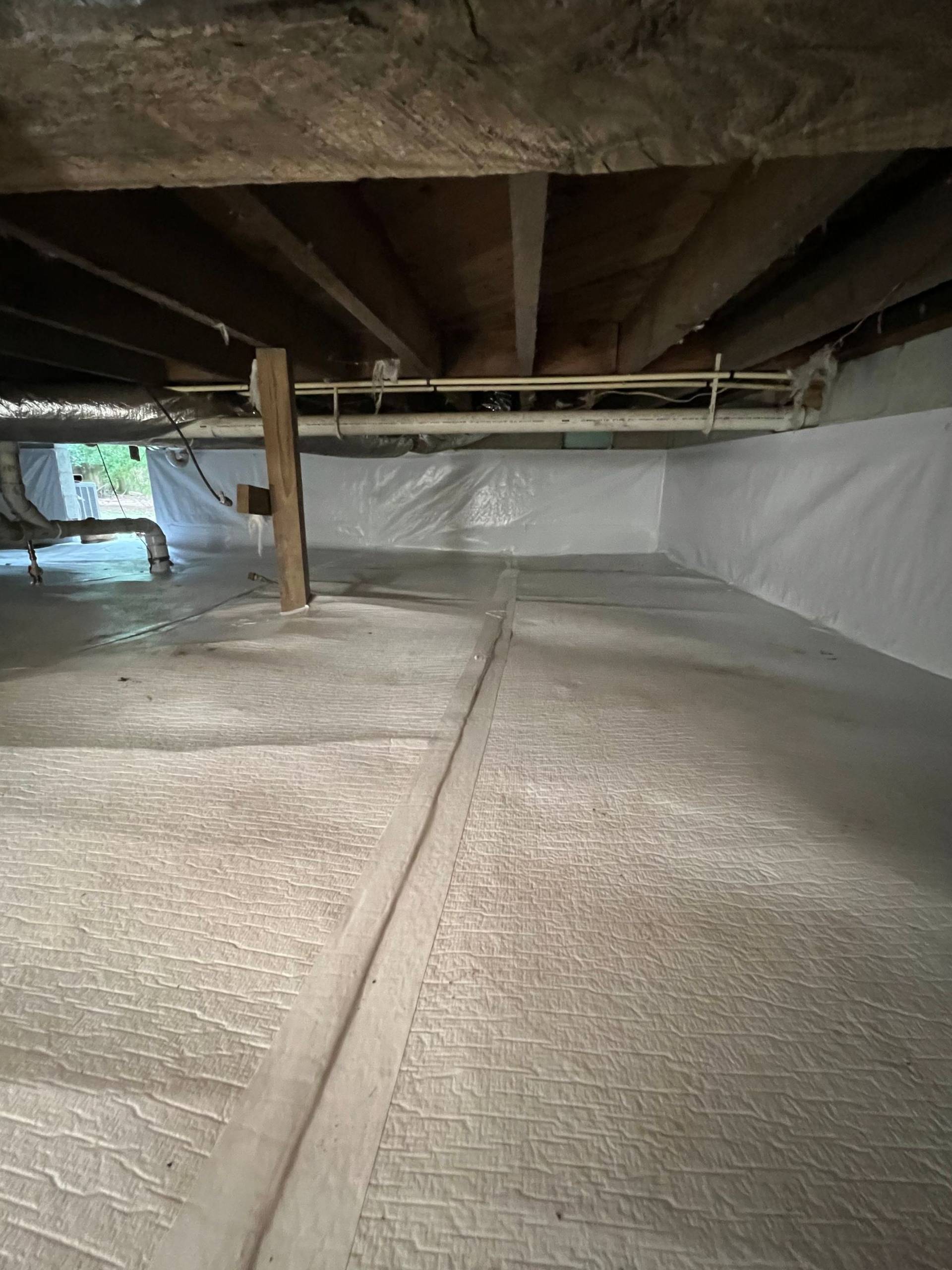 Crawlspace with insulation on walls and floor, beams above, pipes, and wooden support beams.