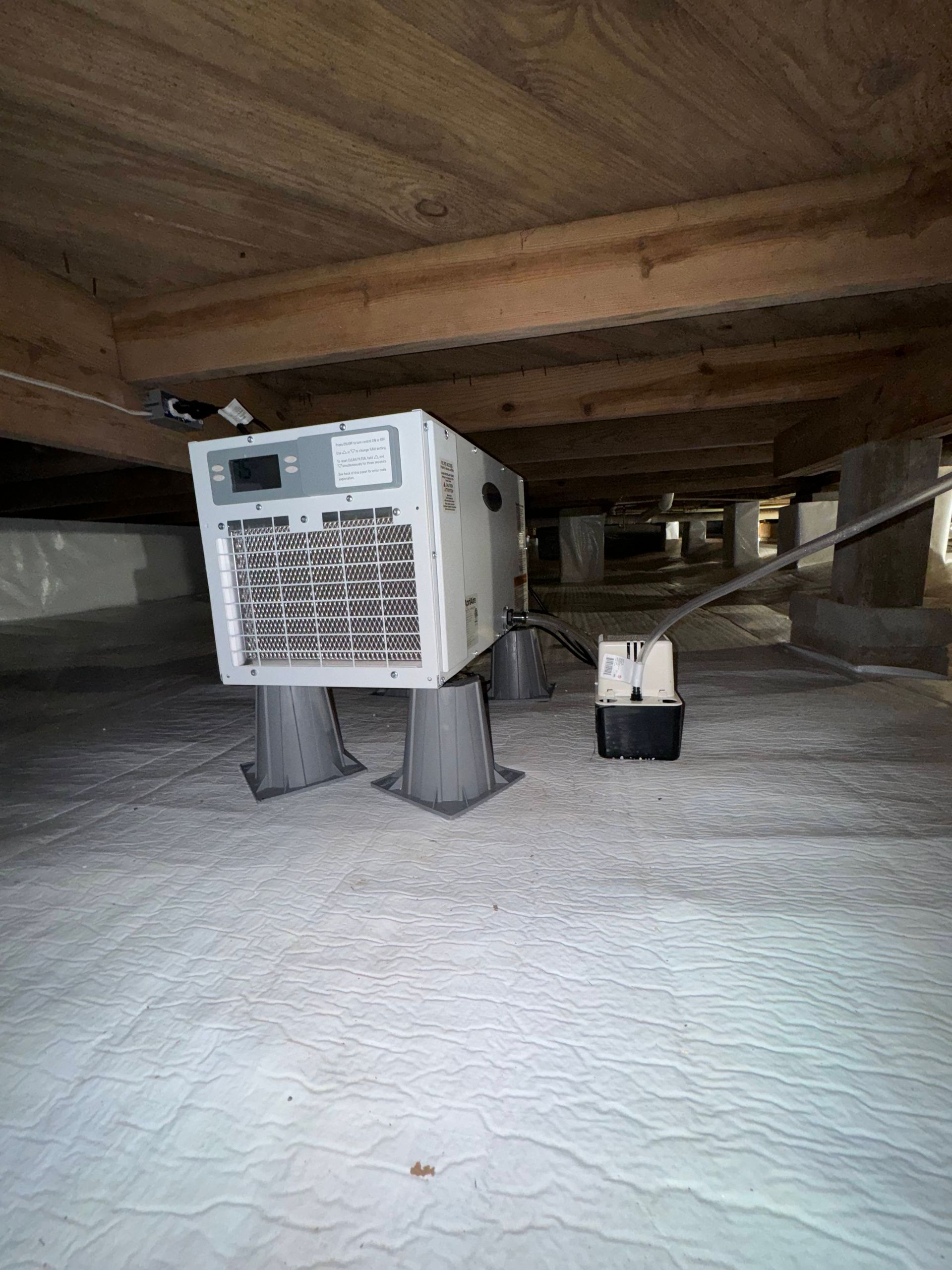 A large white industrial unit under a wooden crawlspace on a concrete floor, with a small bucket beside it.