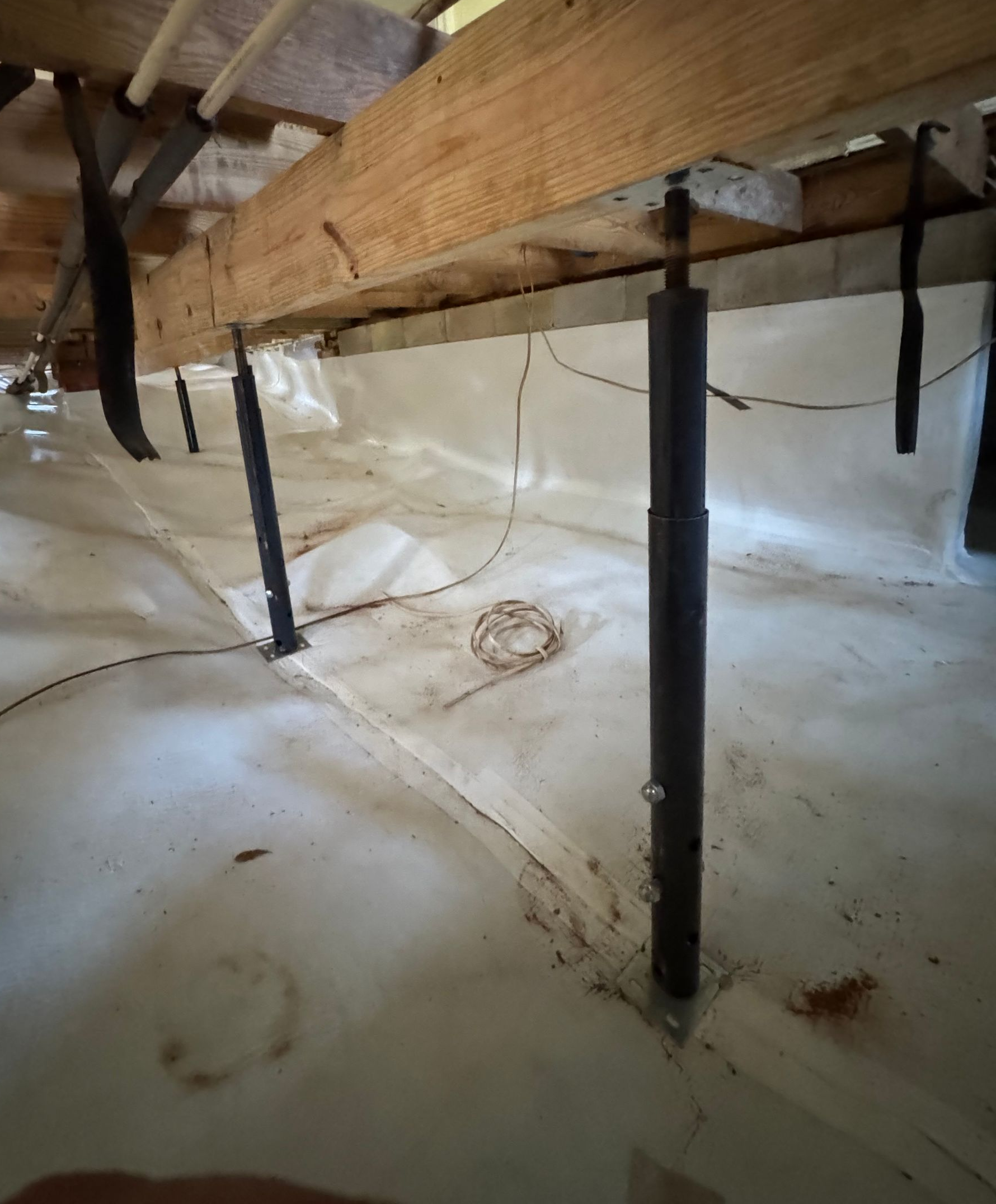 Crawlspace under floor joists with black support posts and white moisture barrier lining the ground.