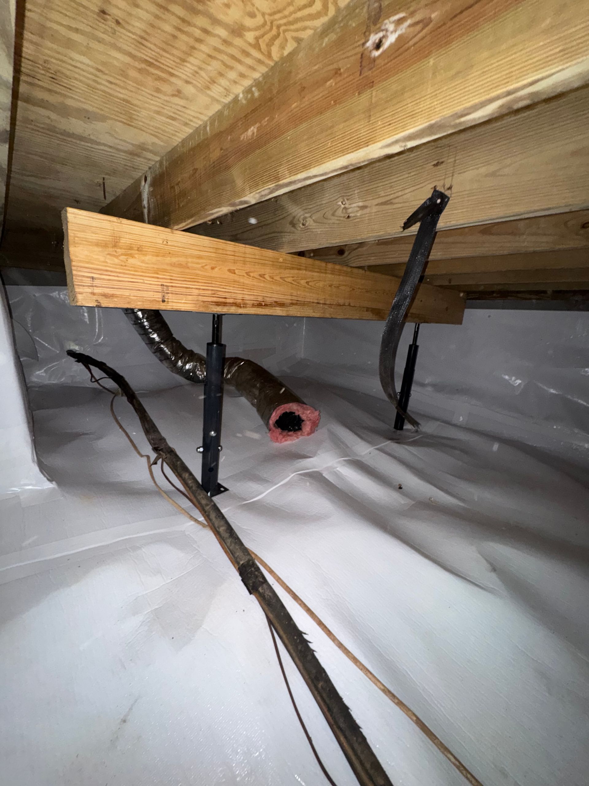 Crawlspace with wooden joists, black wiring, and a red pipe protruding through plastic sheeting.