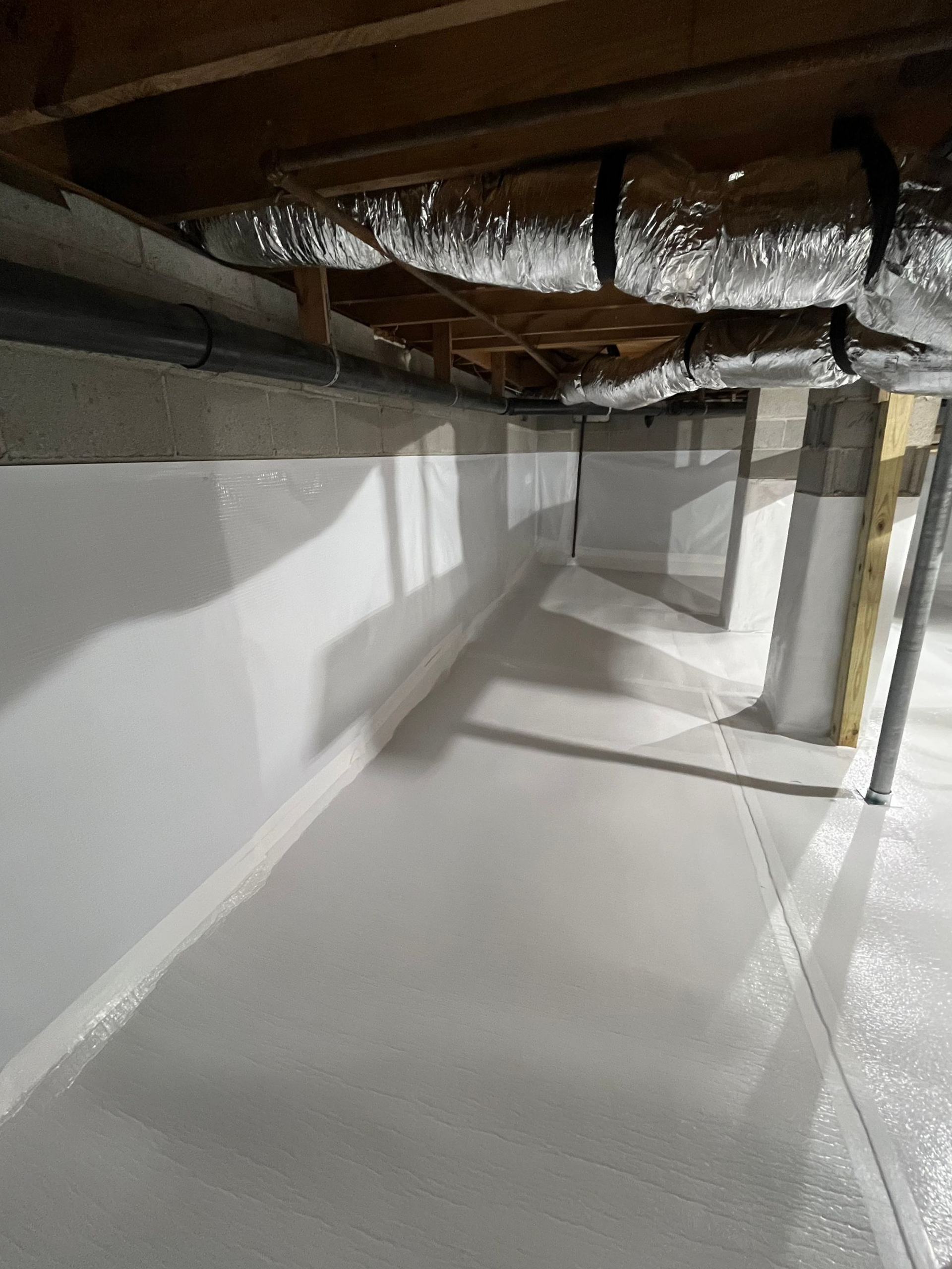 Crawlspace with insulated ducts, white vapor barrier, and support posts in a basement area