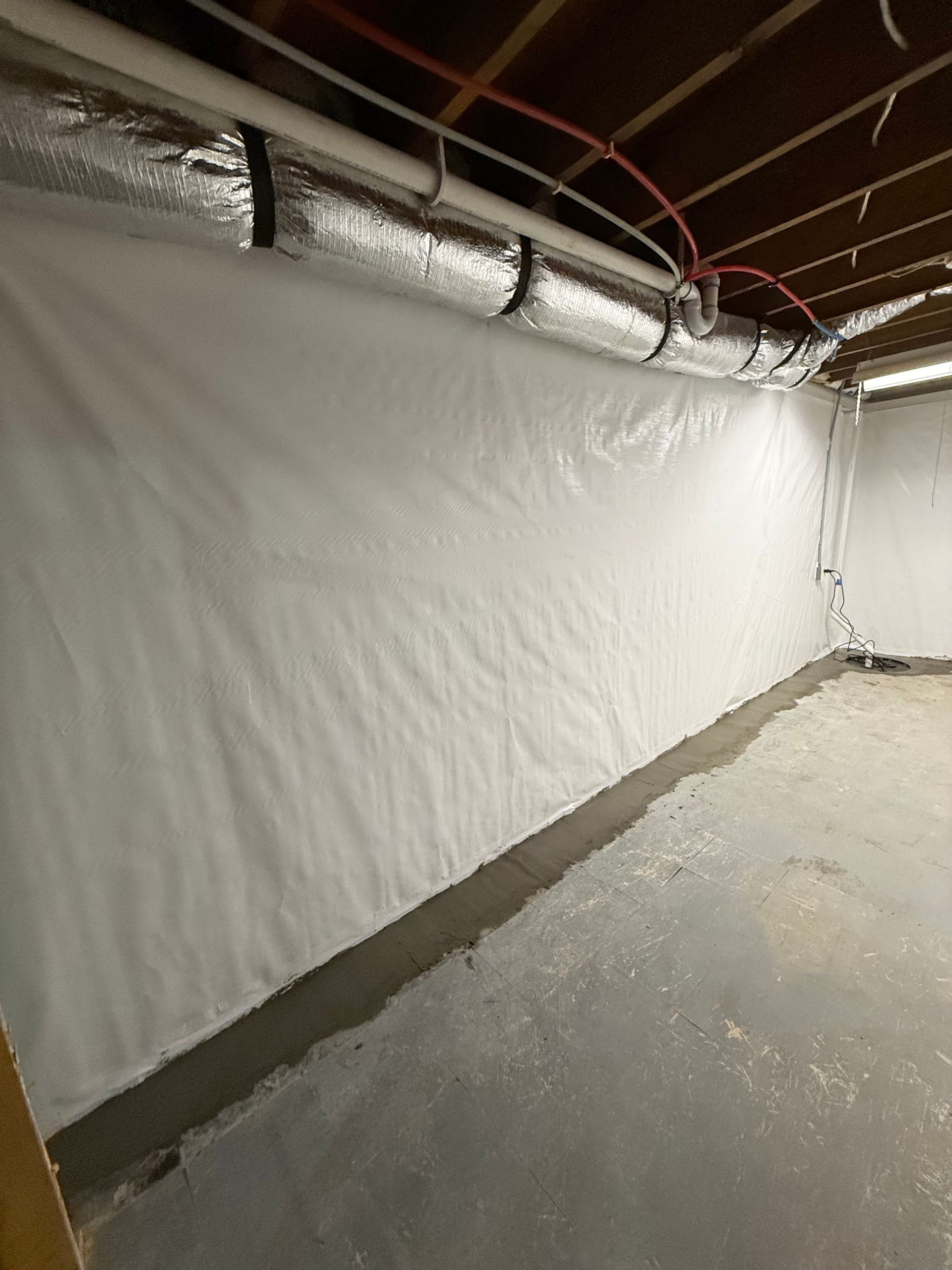 White plastic sheeting covers a basement wall beside an unfinished concrete floor and exposed pipes