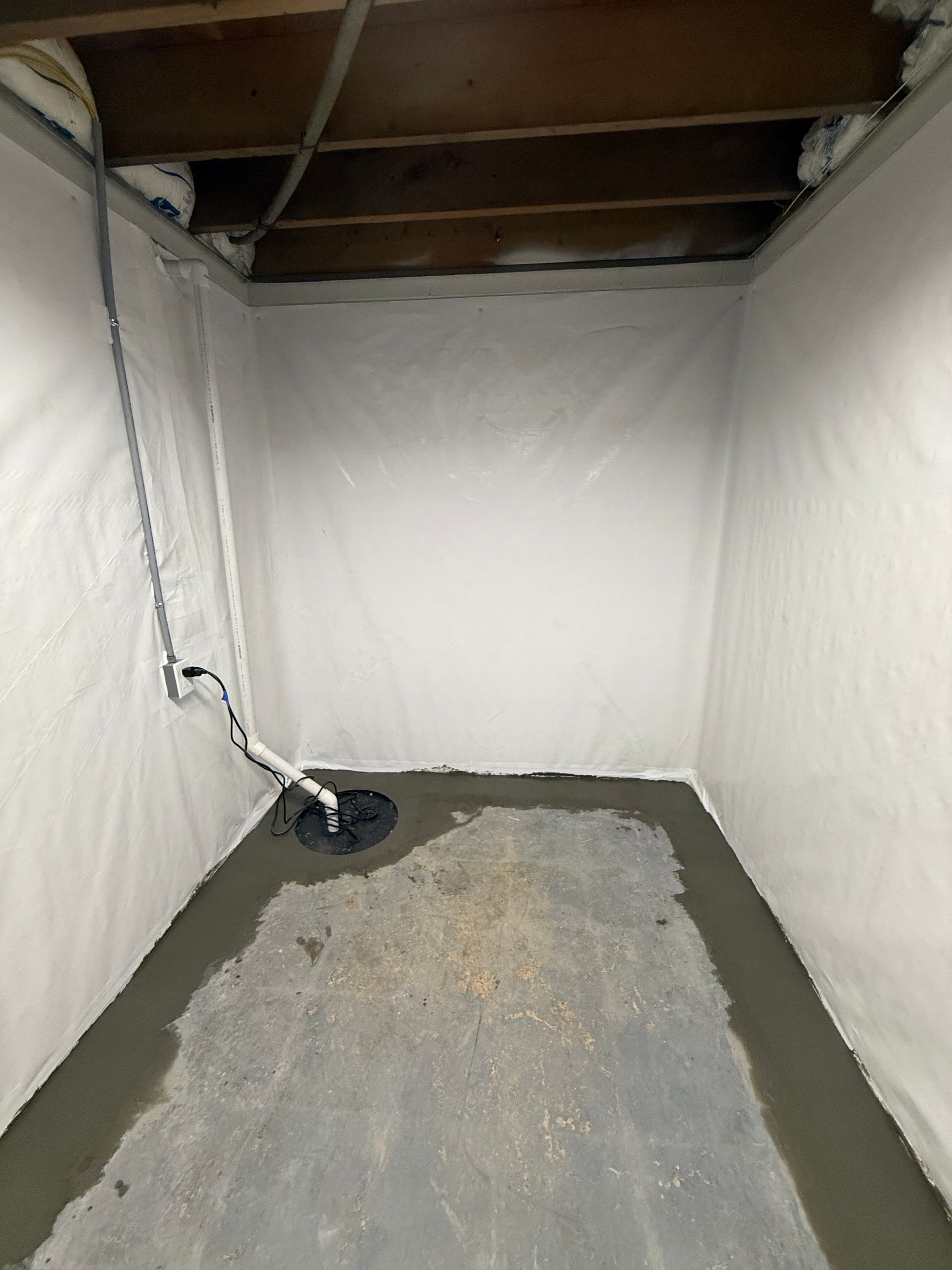 Empty unfinished basement with concrete floor, white walls, exposed ceiling joists, and a sump pump in one corner
