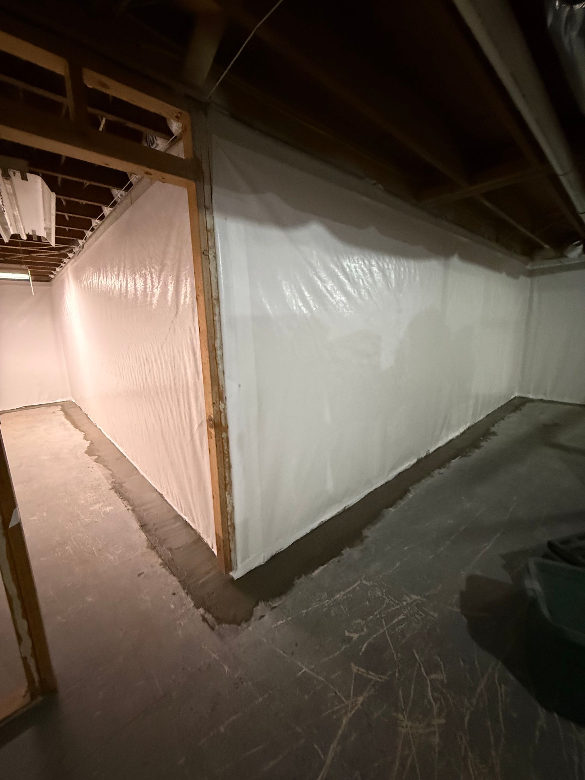Basement hallway with plastic-covered white wall, exposed beams, and dim lighting.
