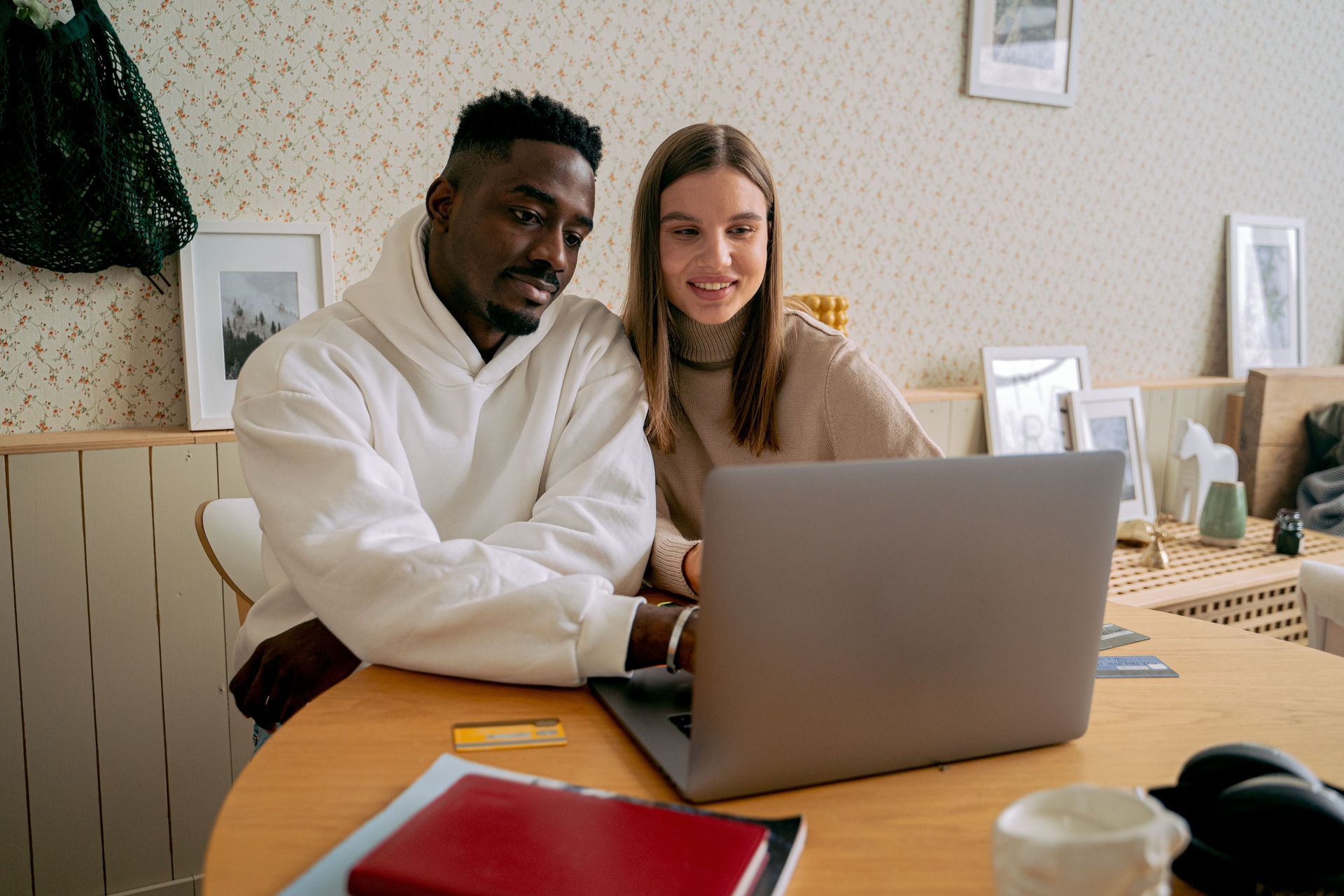 Two people smiling at a laptop at a table in a bright room with papers and a notebook nearby