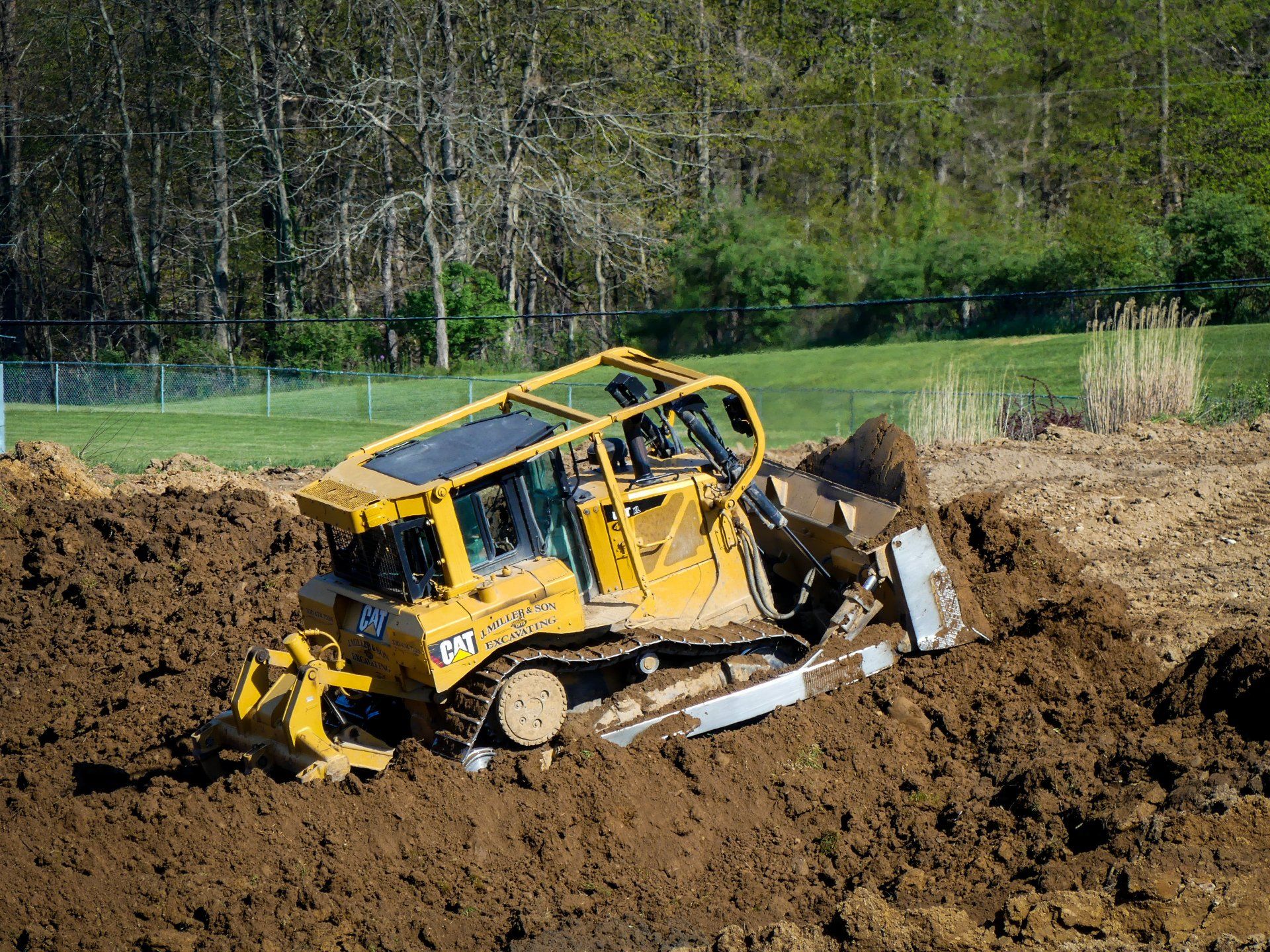 Yellow bulldozer pushing dirt in a muddy field. A forest and fence are in the background.
