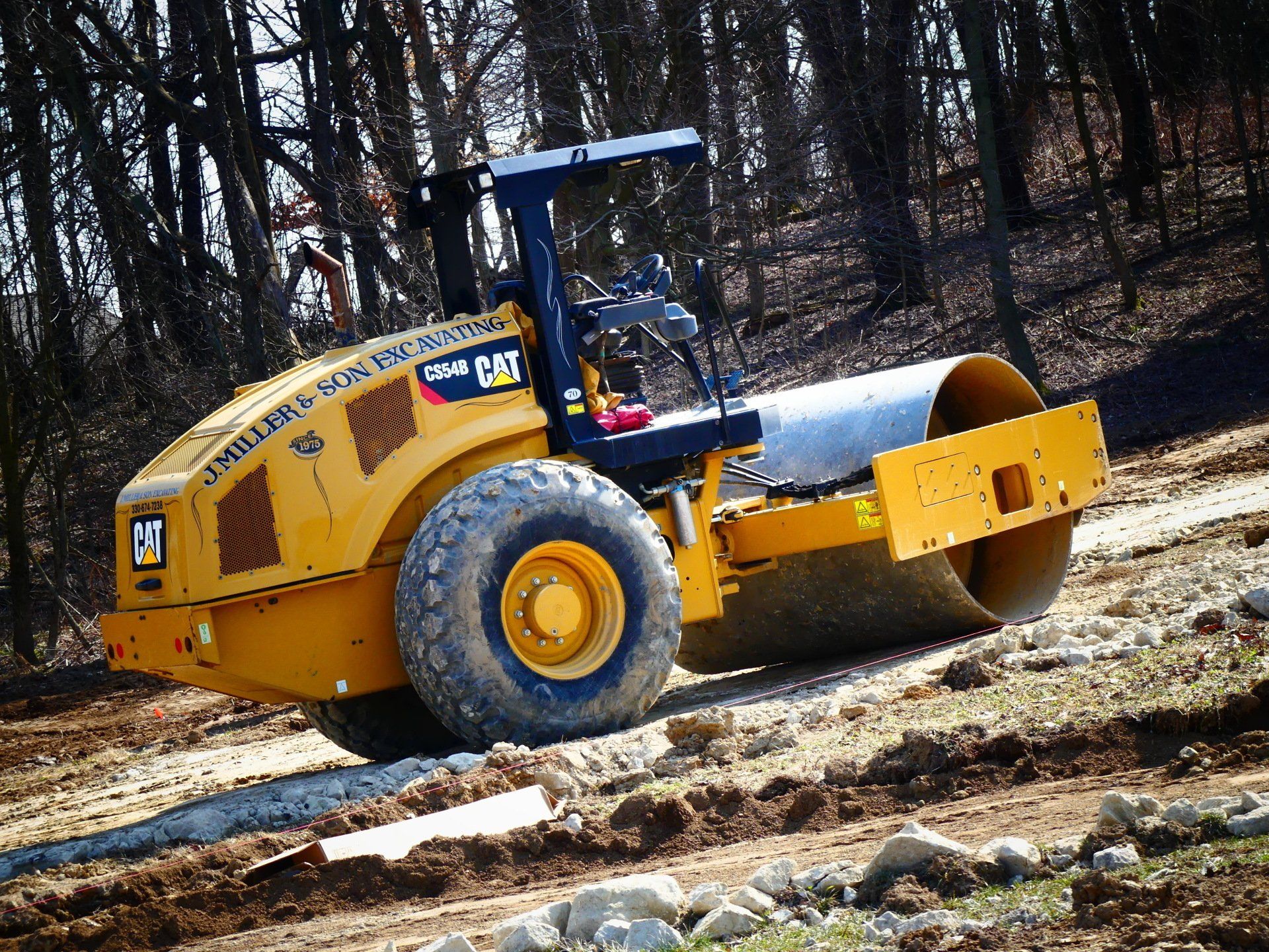 Yellow Caterpillar compactor on a dirt incline, compacting soil near trees.