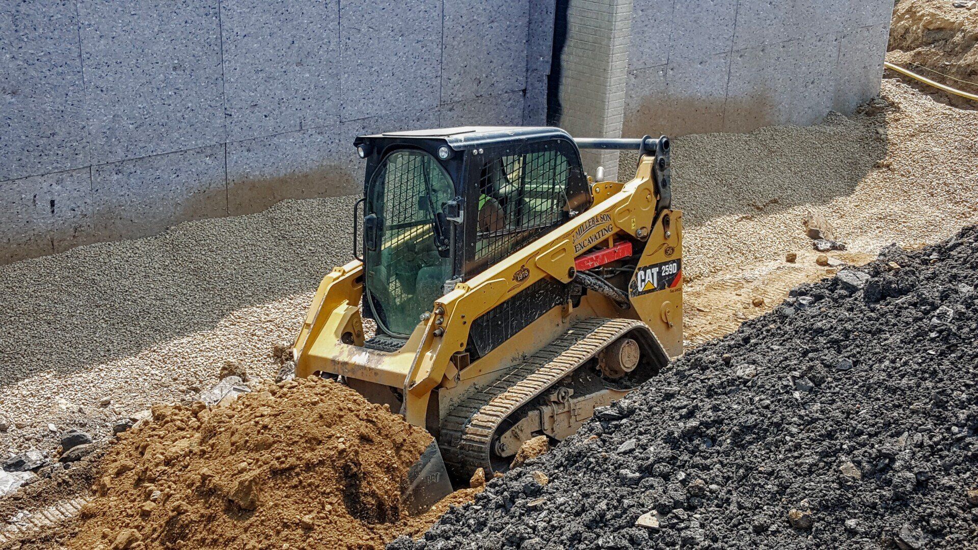 Yellow Caterpillar skid steer digging in a gravel trench next to a concrete wall.