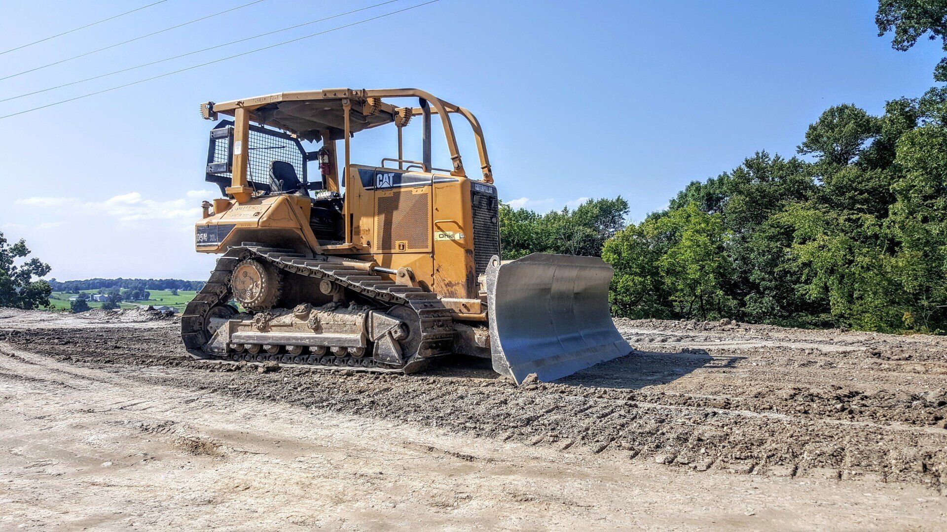 Yellow bulldozer on a gravel road, clearing debris. Sunny day, trees in background.