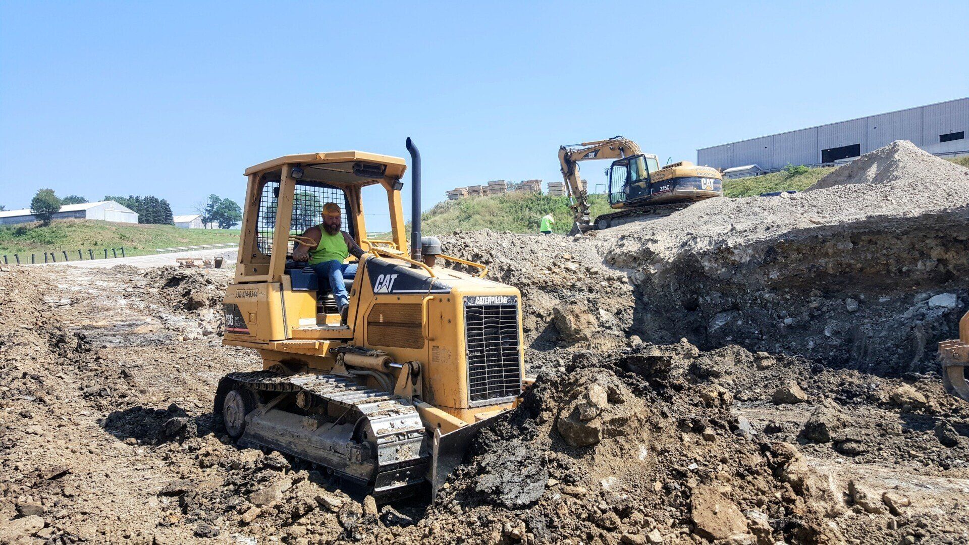 Yellow bulldozer in dirt pit with operator, another excavator in background on a sunny day.