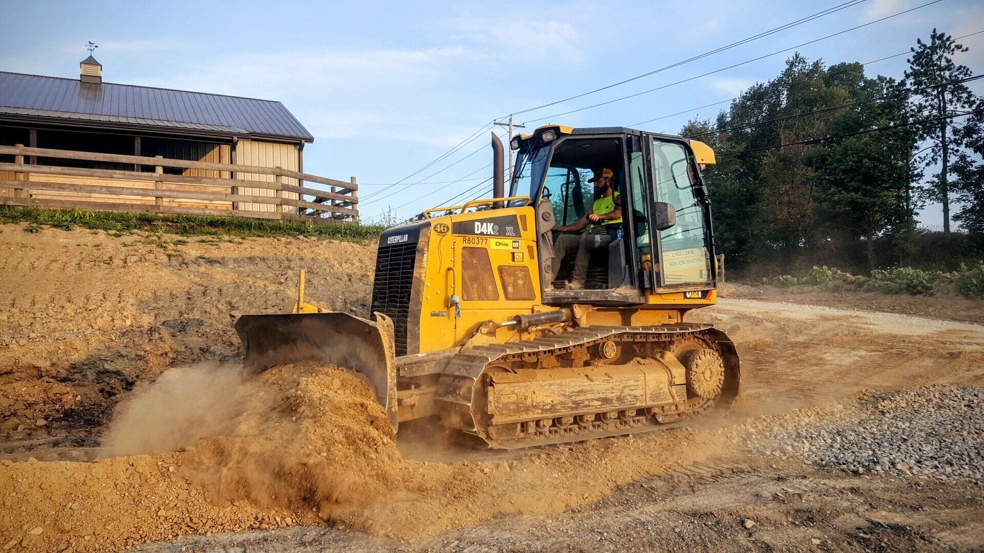Yellow bulldozer grading earth on a construction site. Man in cab, barn in background.