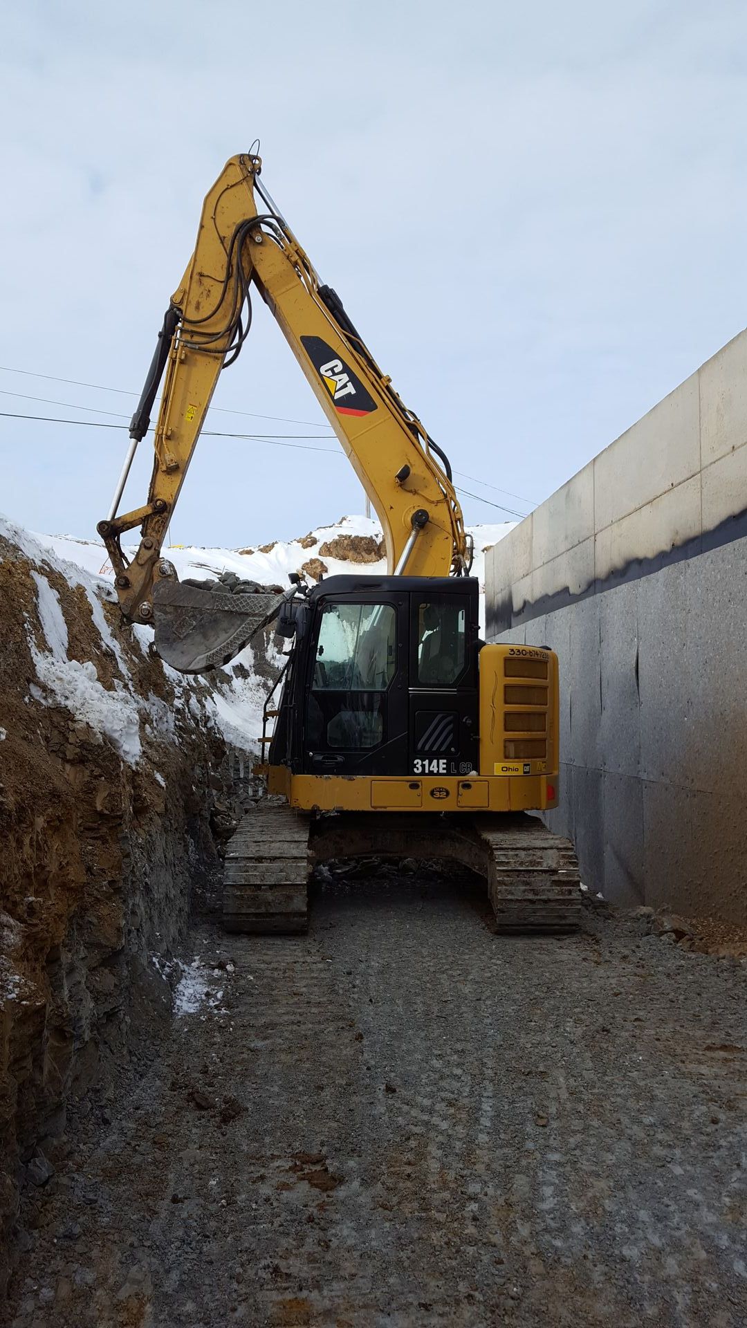 Yellow Caterpillar excavator digging earth near a concrete wall. Snowy background.