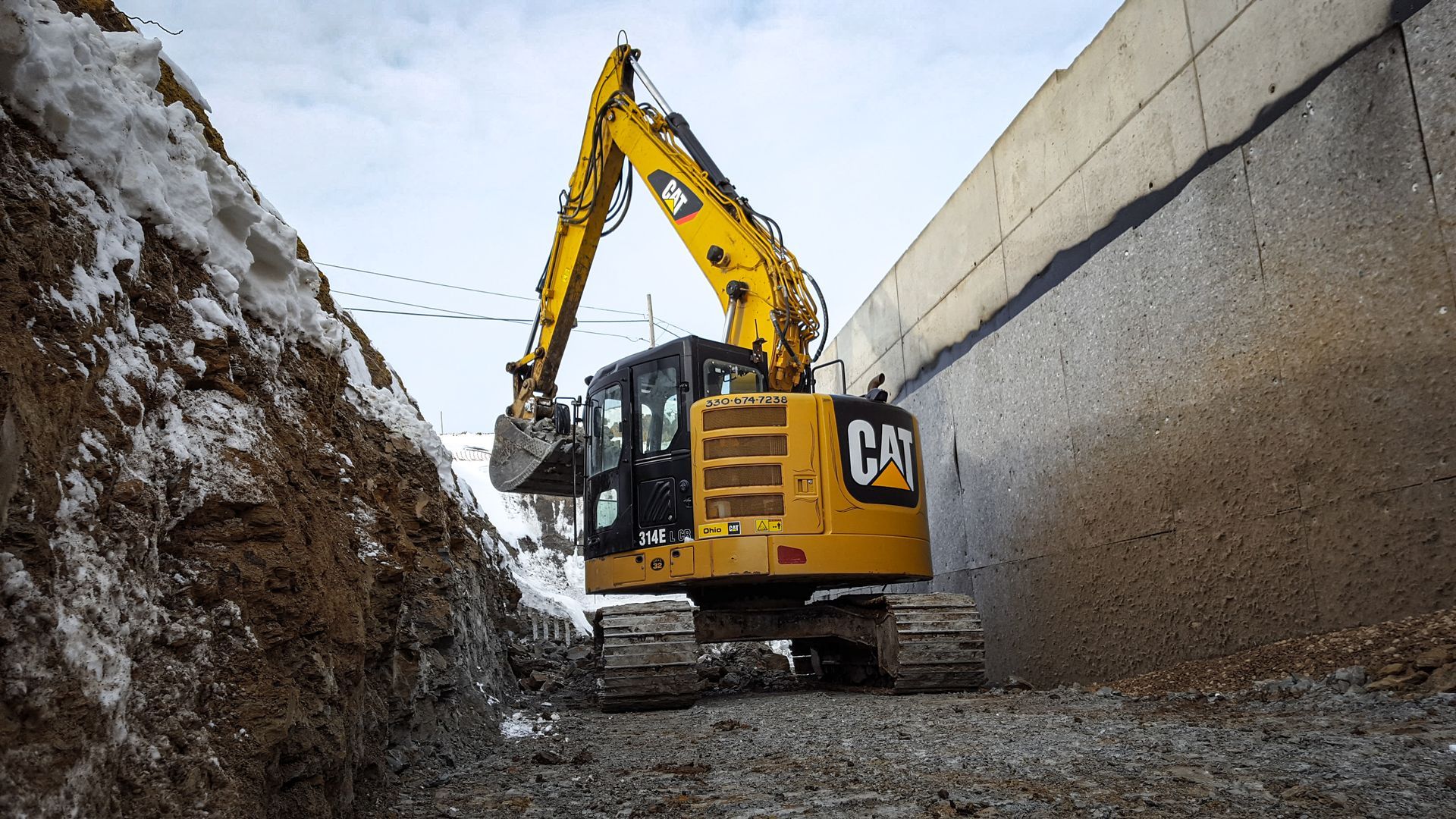 Yellow Caterpillar excavator digging in a trench between earth and concrete wall.