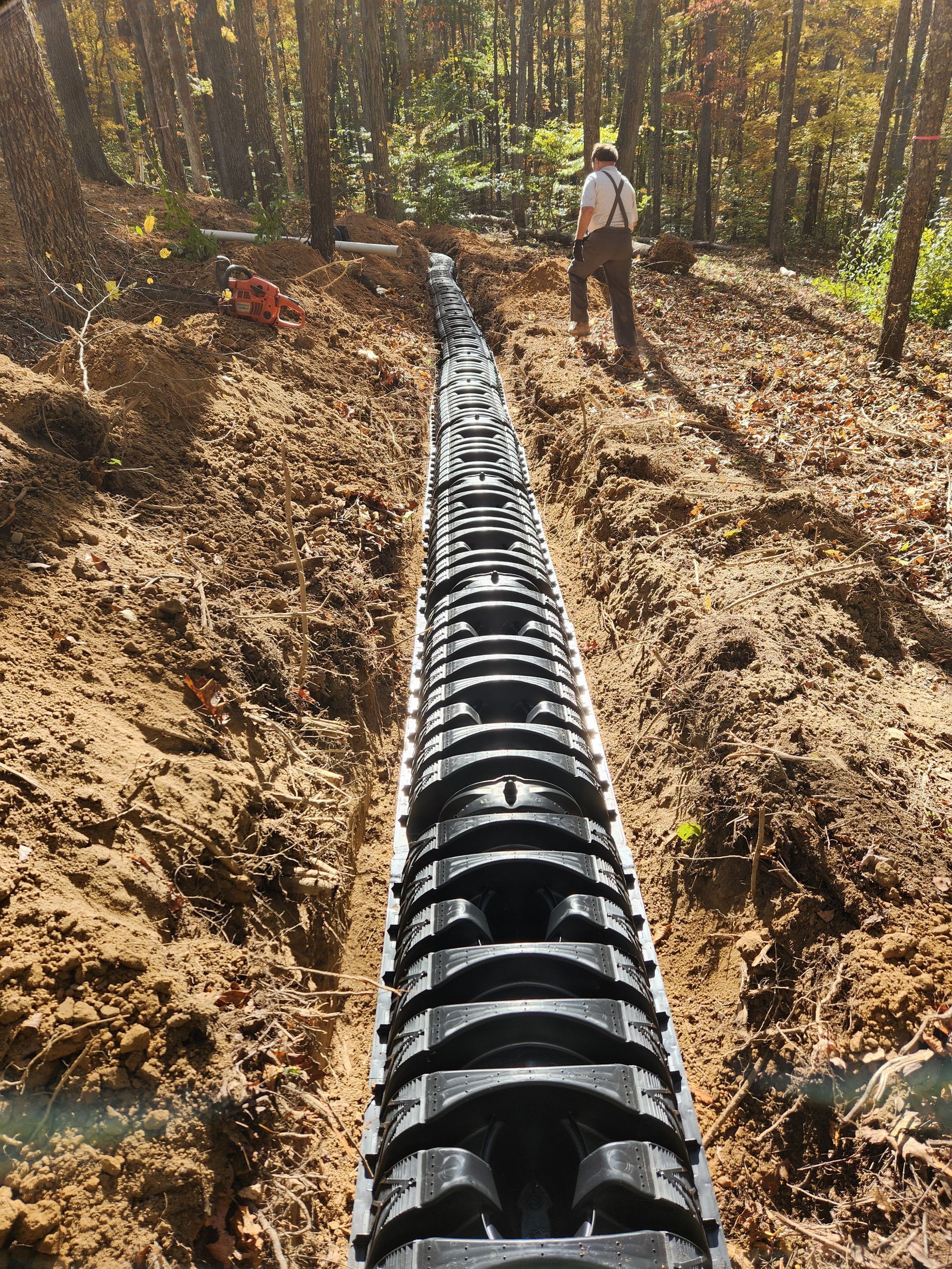 Long trench with black drainage system installed; person standing at end in wooded area.