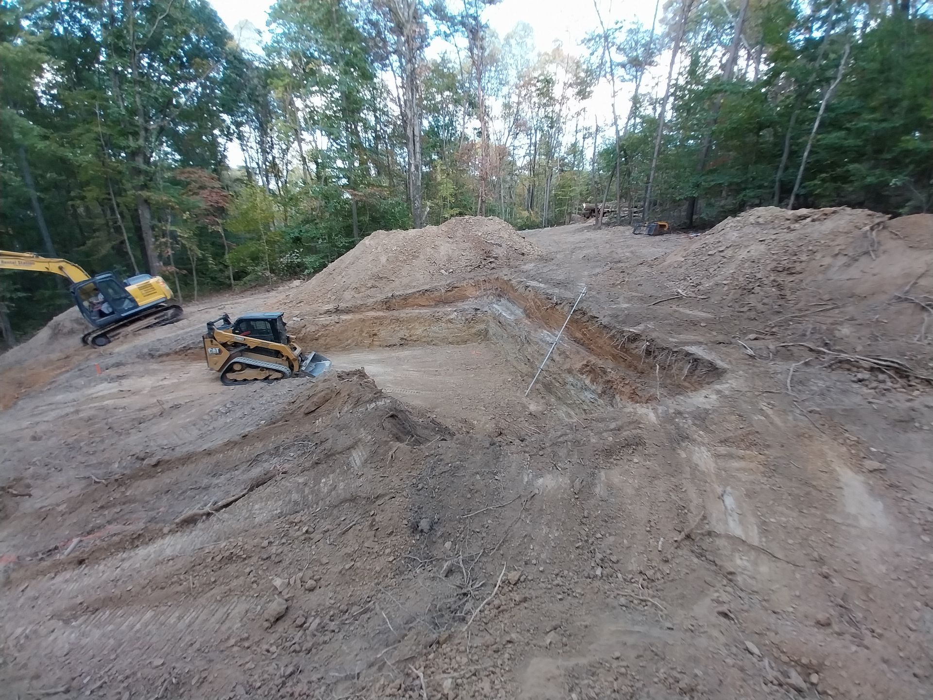 Construction site: two bulldozers and an excavator moving dirt, surrounded by trees.