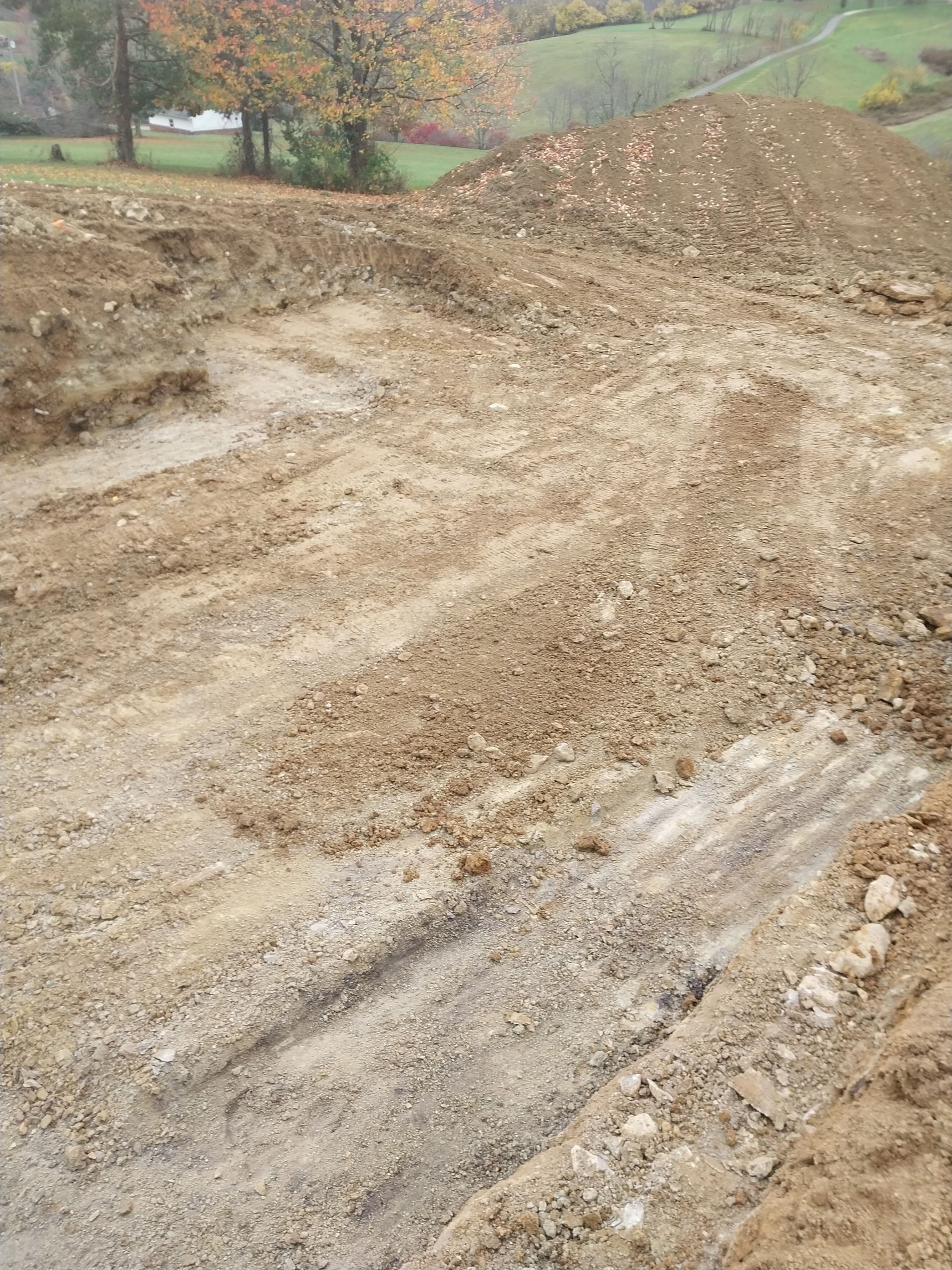 Construction site with dirt piles and tire tracks in the mud, trees and a distant hill.