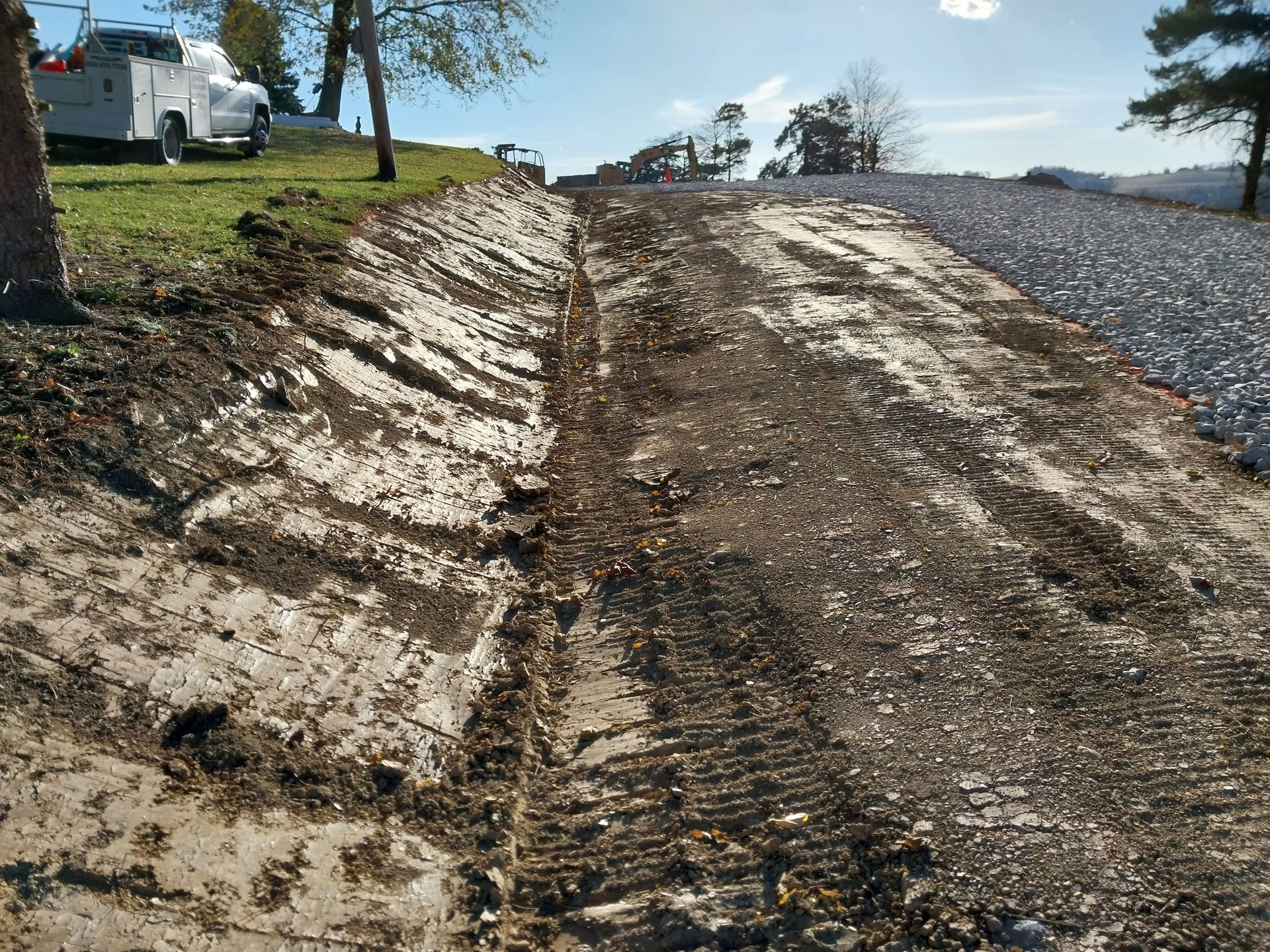 Dirt and gravel pathway construction, with machinery, near a grassy hill and trees.