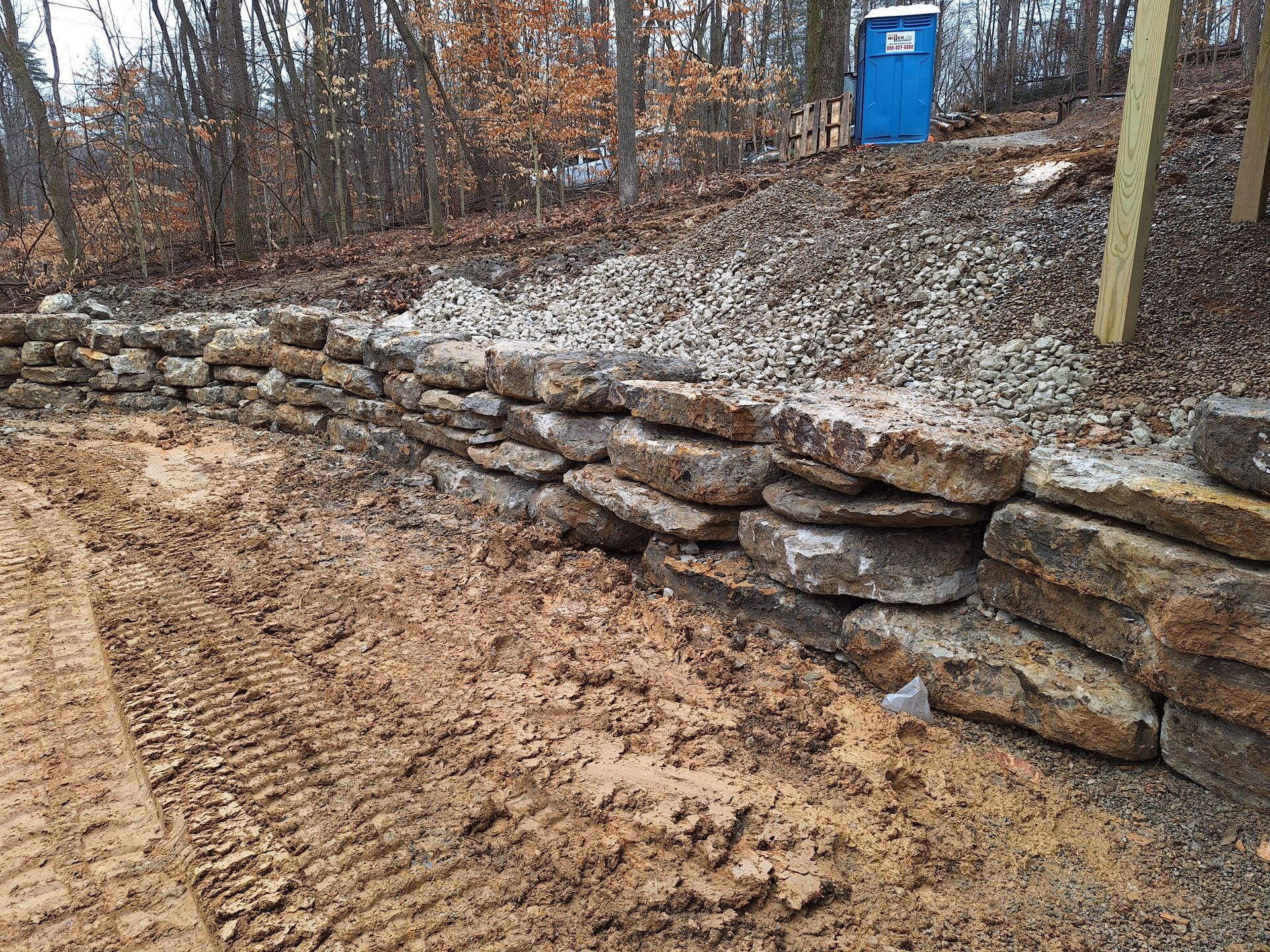 Stone retaining wall next to muddy ground in a wooded area with a blue porta-potty in the background.