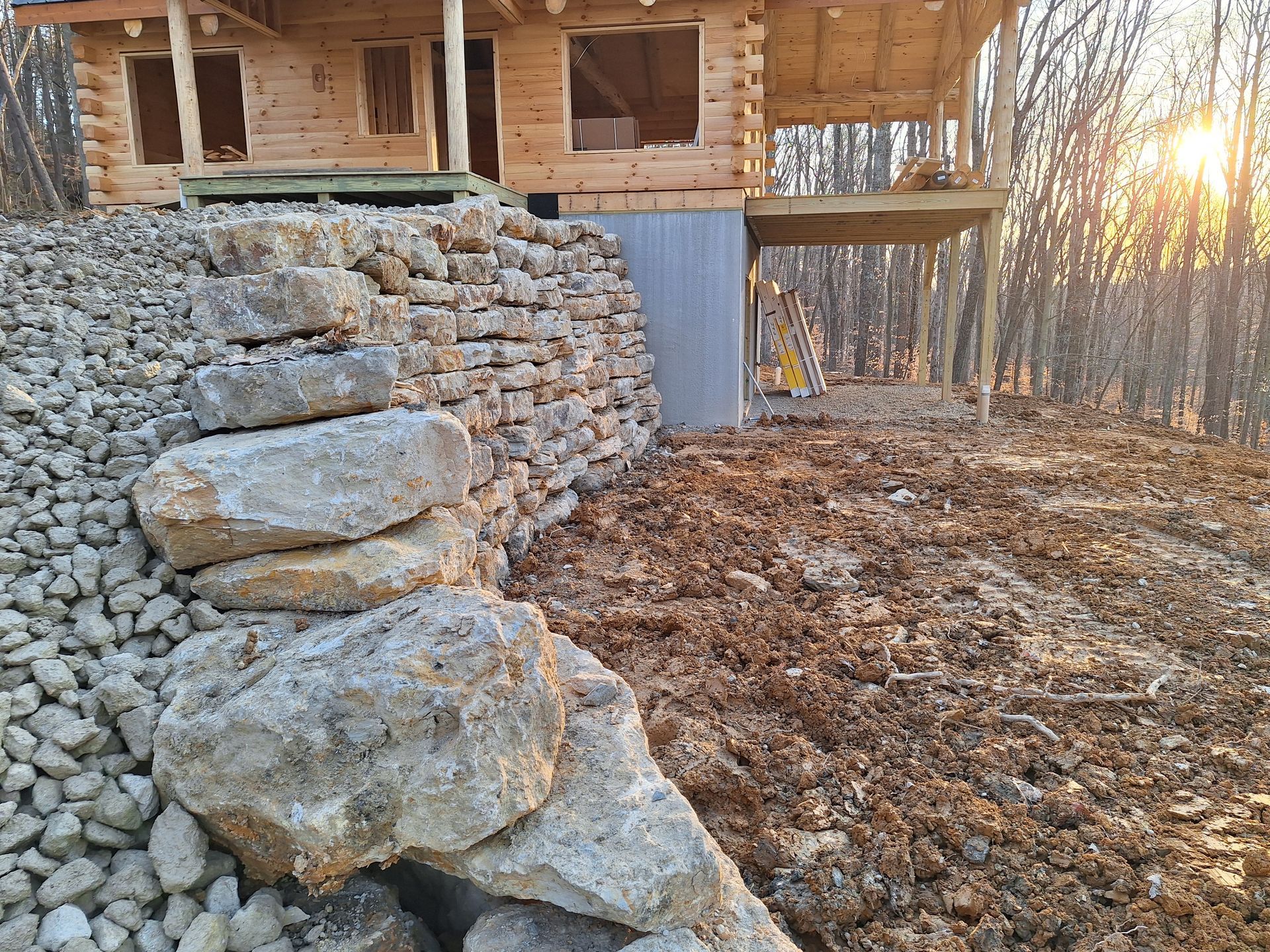 Stone steps lead to a log cabin under construction. The structure sits on a hillside with dry leaves and trees.