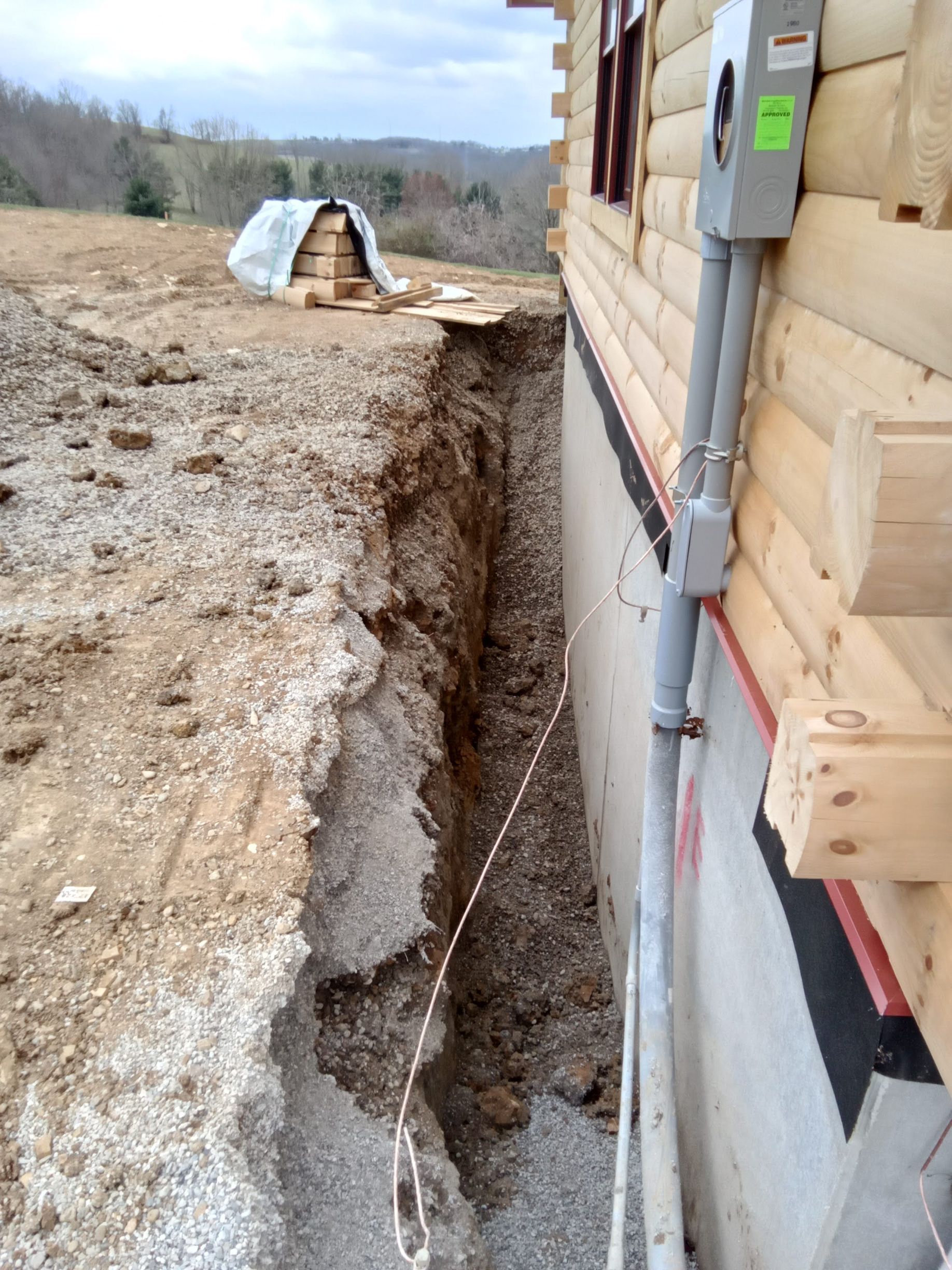 A deep trench next to a log cabin under construction; electrical conduit on the wall, pile of wood in the background.