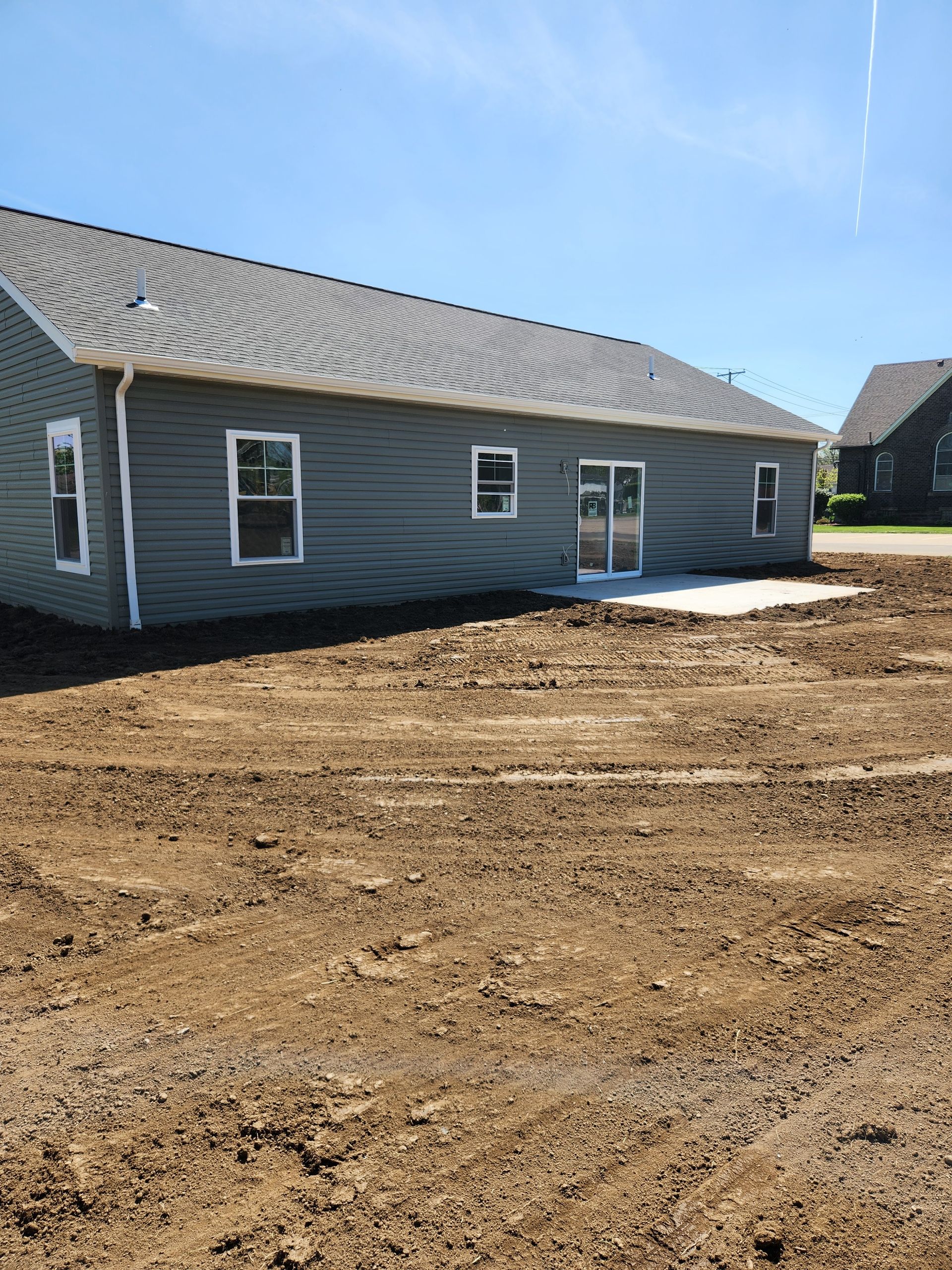Back of a gray house with a concrete patio; bare dirt in the yard.