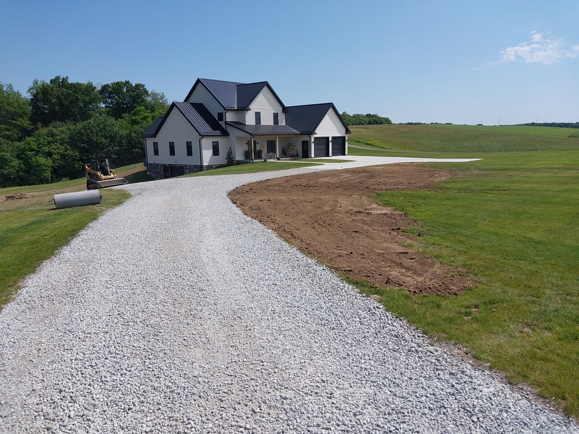 Gravel driveway leading to a two-story white house with a black roof and two-car garage, on a grassy field.