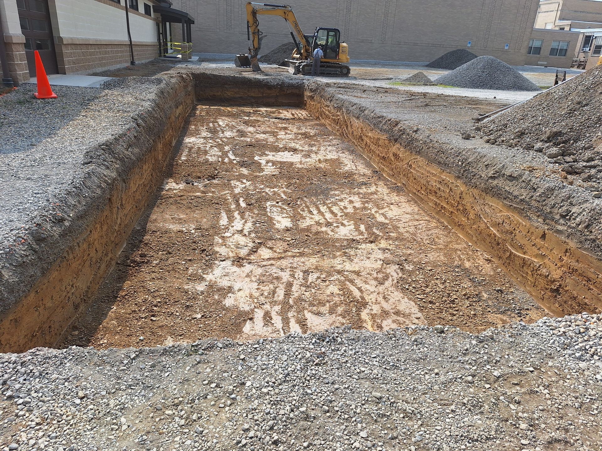 Construction site: Excavated rectangular pit, backhoe, piles of gravel, orange cone.