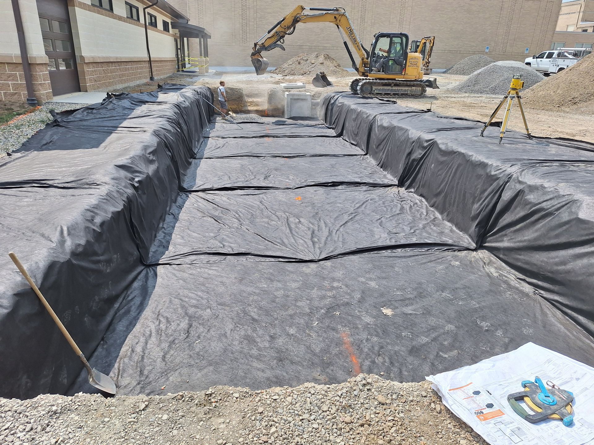 Construction site with black lining in trenches; excavator in background, blueprints in foreground.