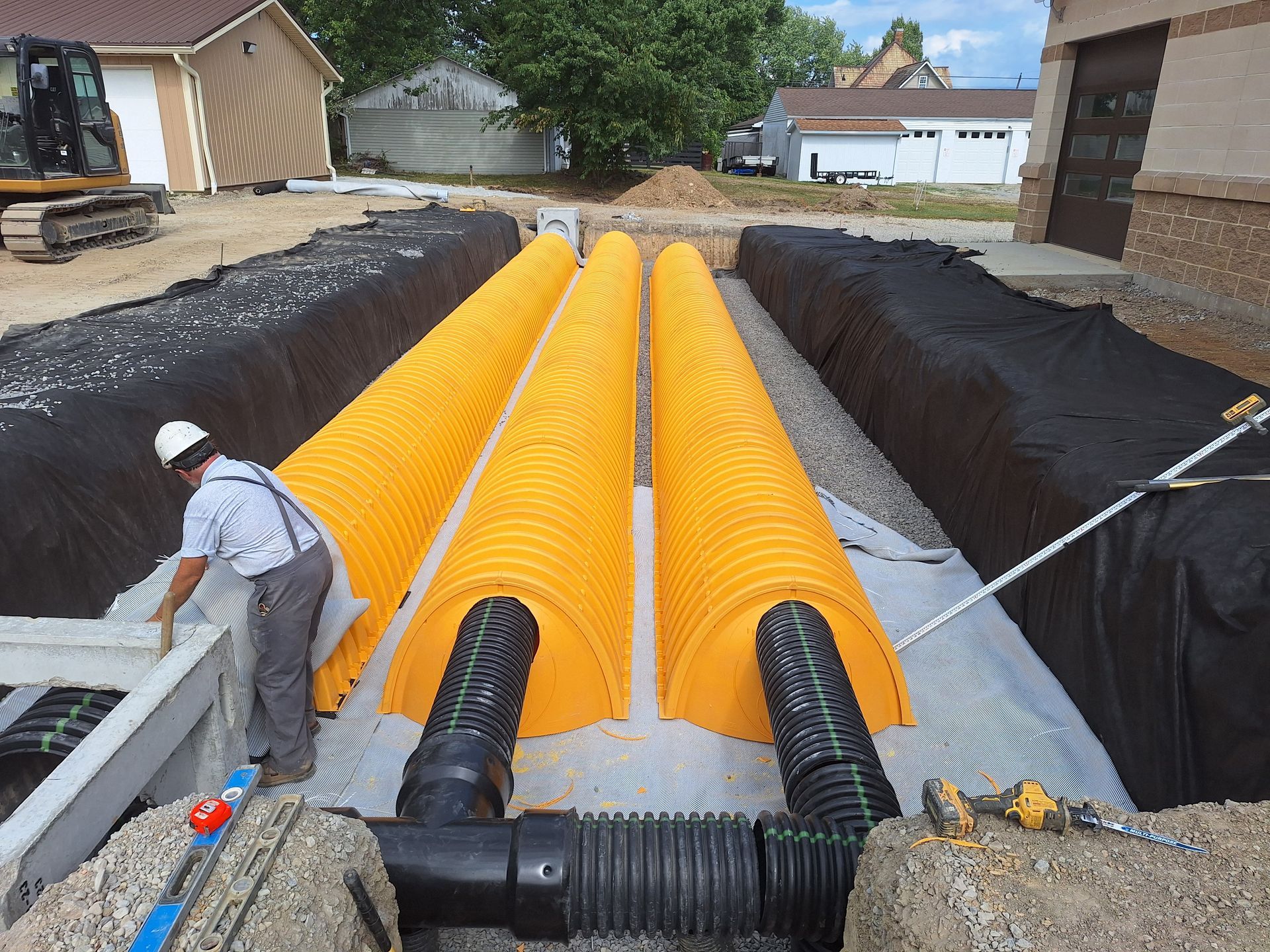 Construction site with yellow drainage pipes, lined with black fabric, and a worker inspecting the setup.