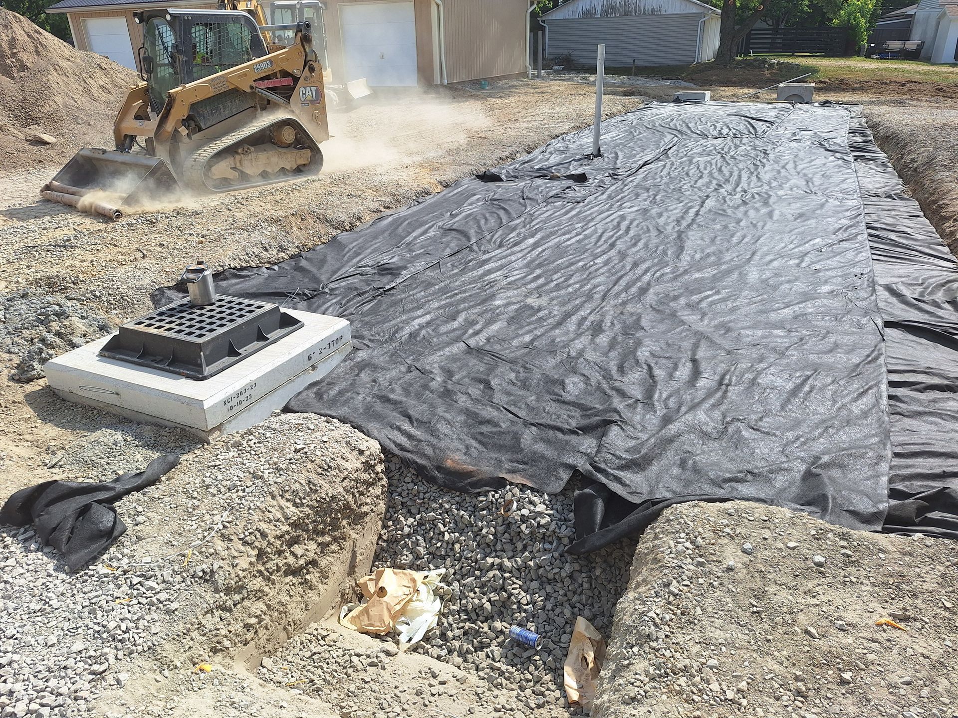 Construction site with excavator and drainage system components; black fabric covers gravel.