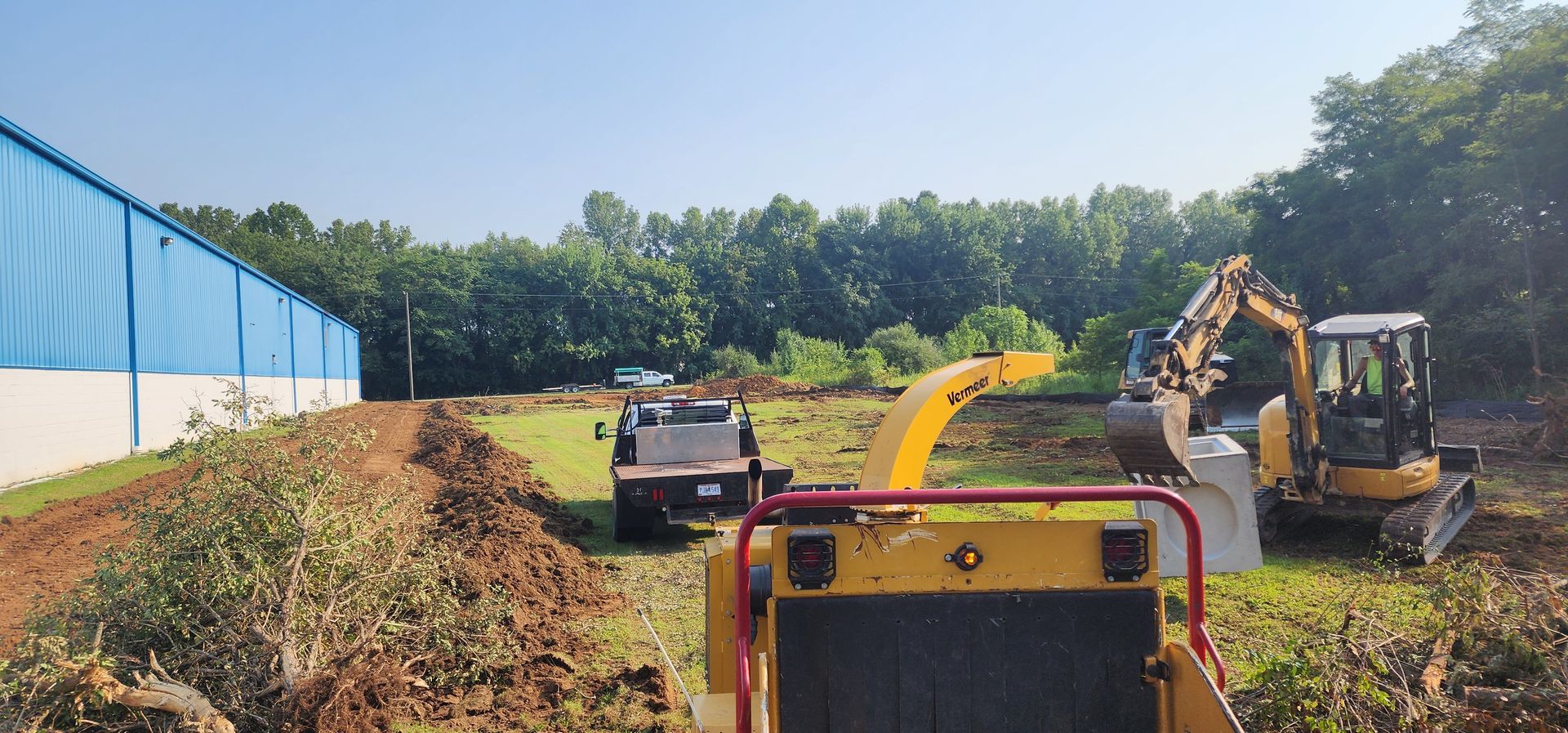An excavator feeds a wood chipper. A truck and building are in the background on a sunny day.