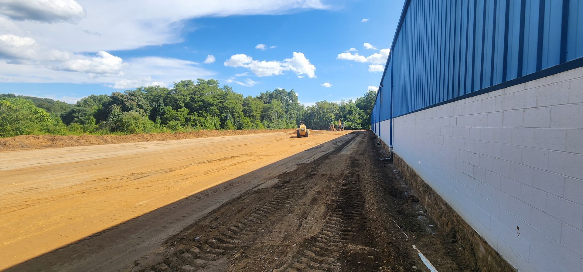 A construction site next to a blue and white building, with earthworks and trees in the distance under a blue sky.