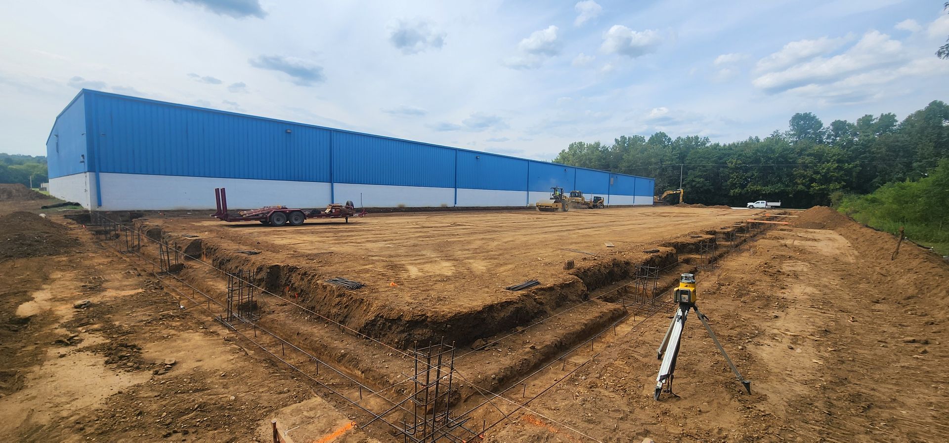 Construction site: long blue building, excavated dirt, trees, and cloudy sky.