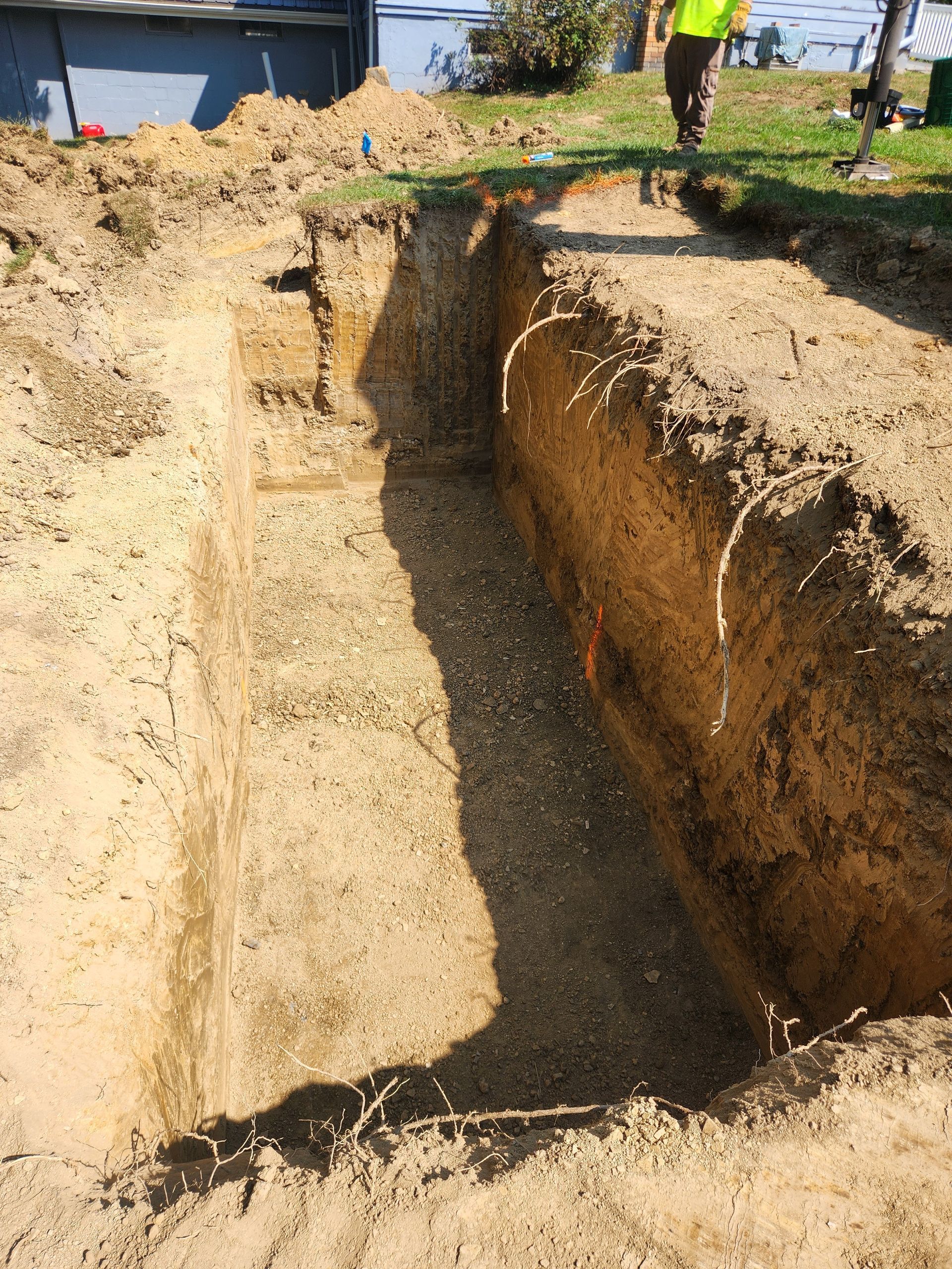 Trench filled with gravel, freshly dug in a grassy area with a worker in the background.