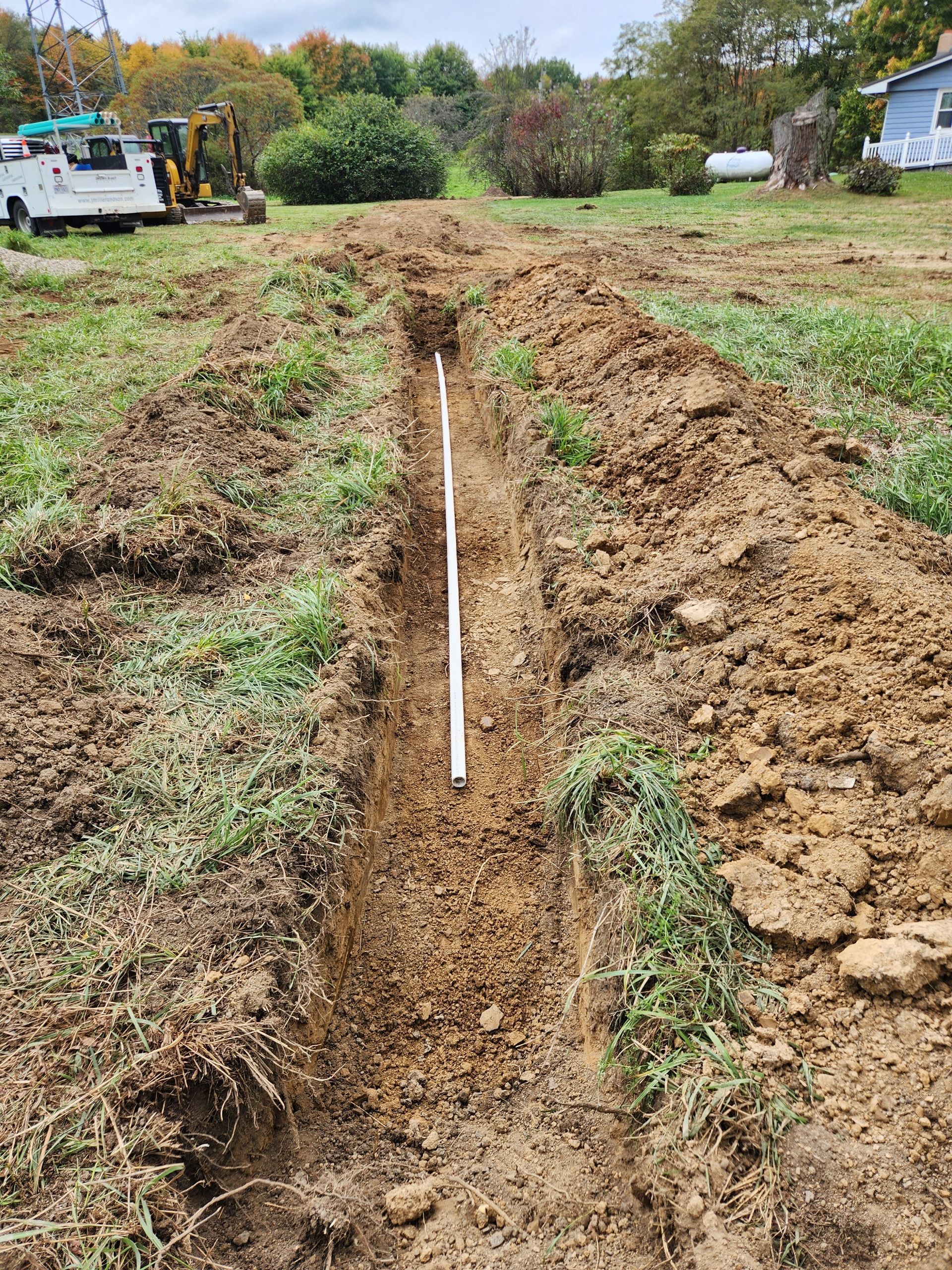 Trench with white pipe laid in it, dug in a grassy field, construction vehicles visible in background.